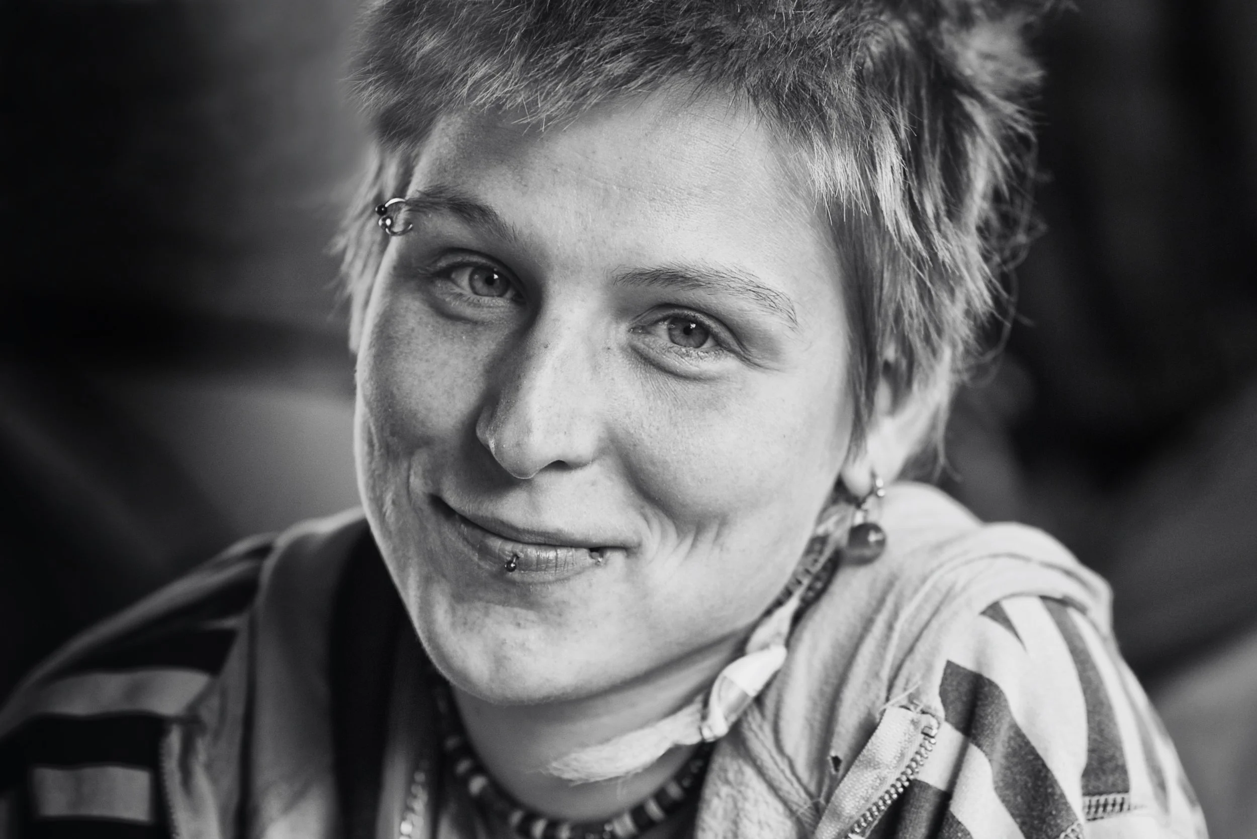Schwarz-weiß Portrait einer smiling jungen Frau mit kurzen, stacheligem Haar und Piercings. Sie trägt Armbänder, Ohrringe und ein kariertes Hemd.