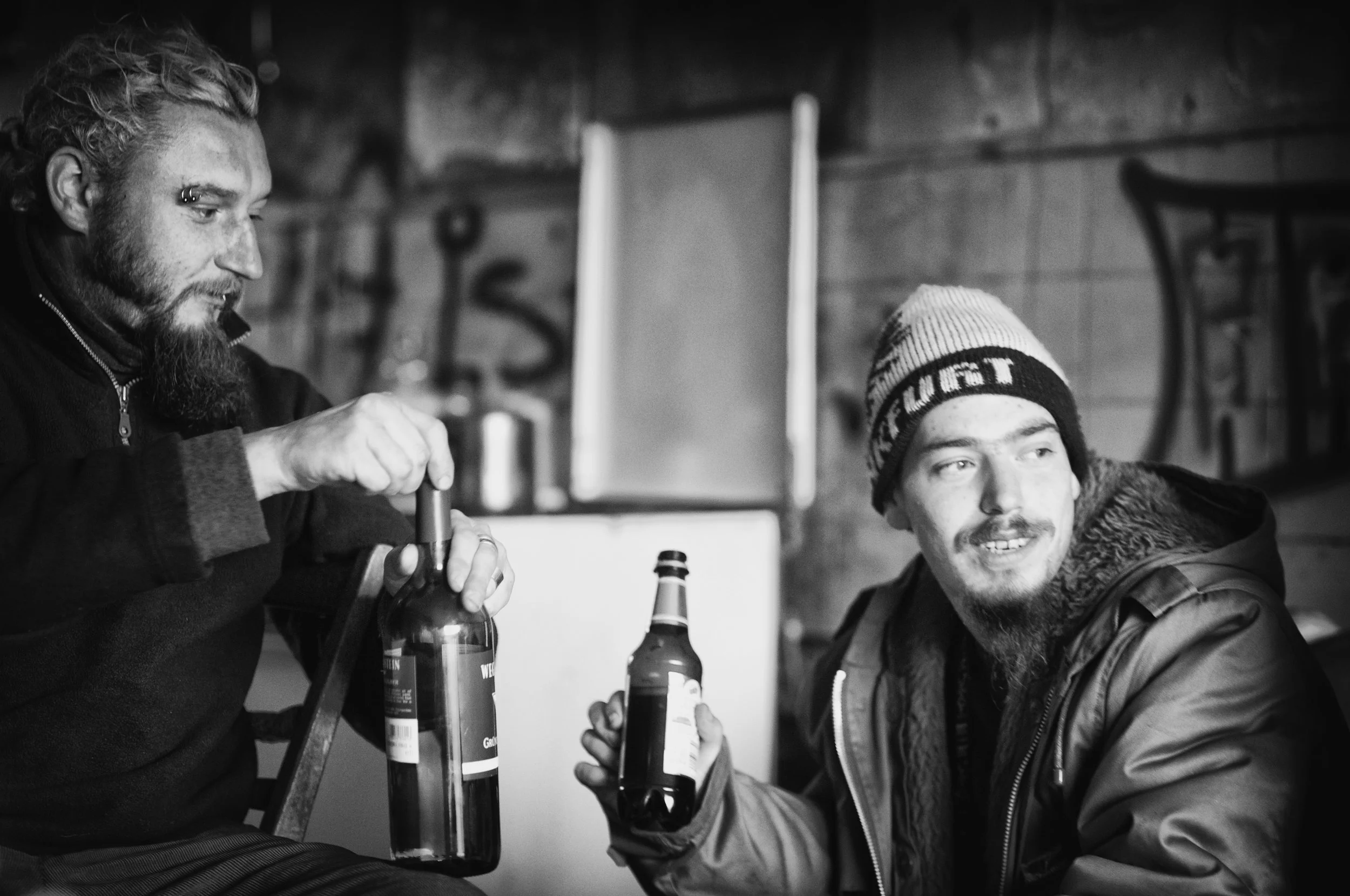 Zwei Männer in einer Holzbar, einer öffnet eine Weinflasche, der andere hält eine Bierflasche, beide lächeln und genießen ihre Getränke.