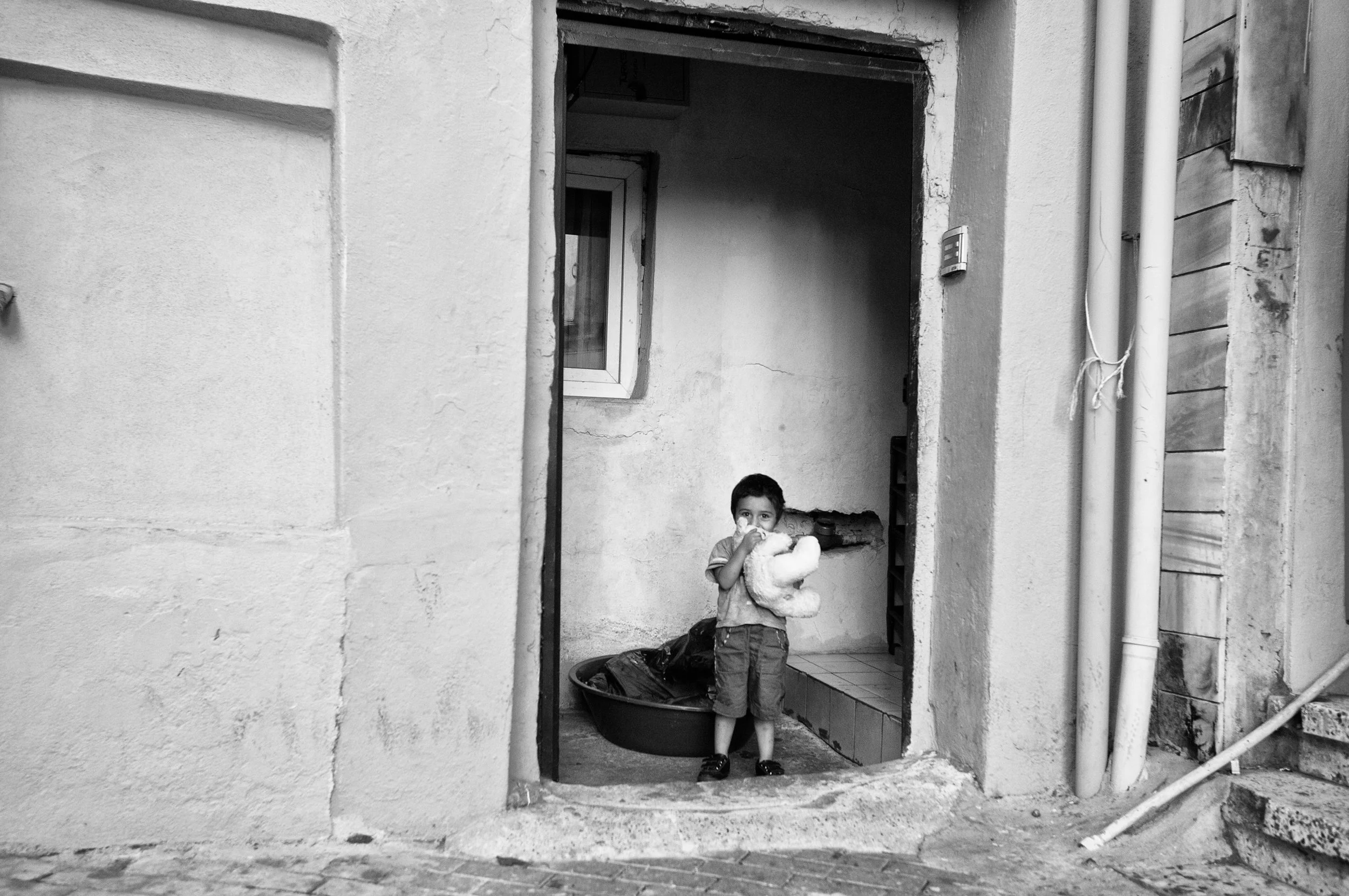 Ein Junge steht in einer Türöffnung, hält ein Kuscheltier und schaut in die Kamera, im Hintergrund eine Wand mit einem kleinen Fenster.