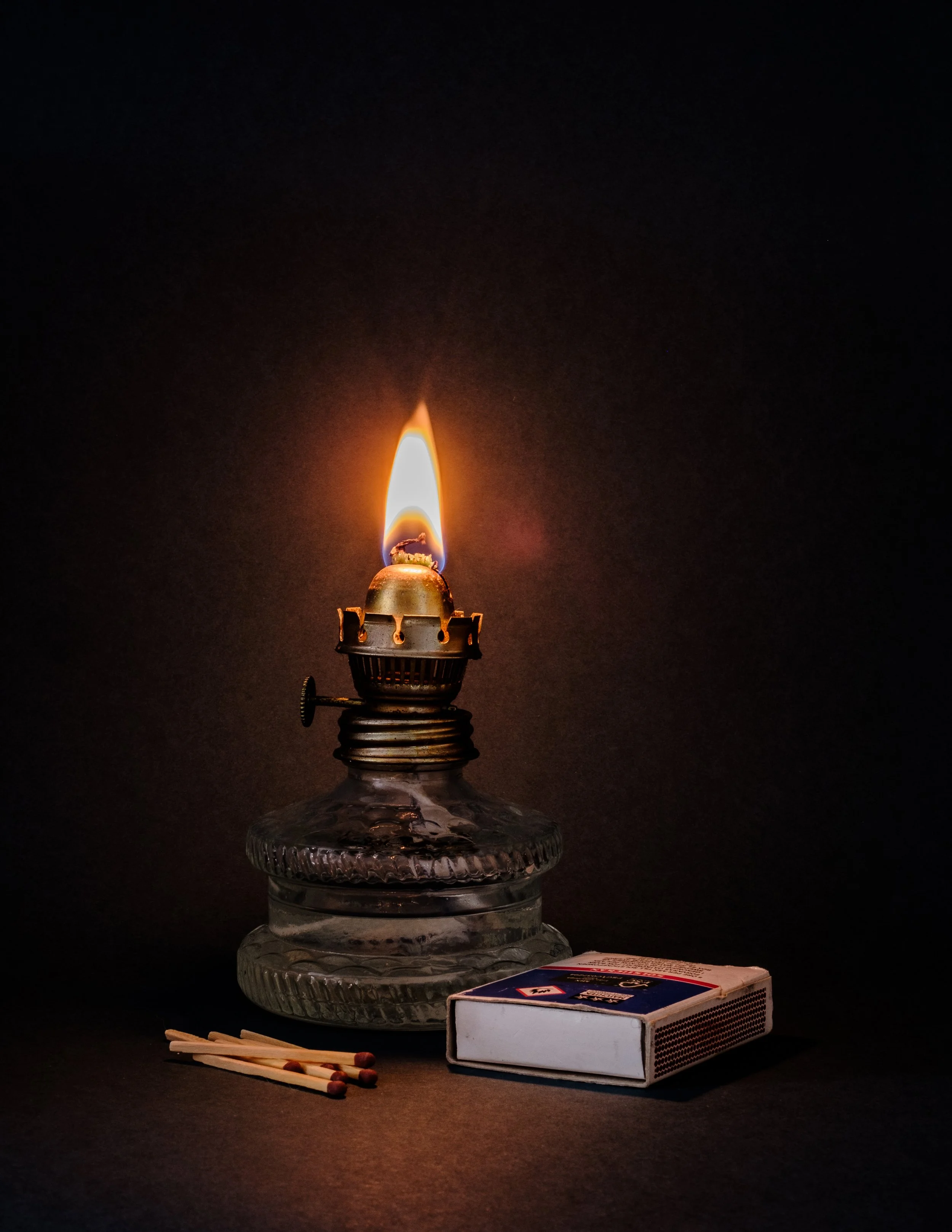 Eine alte Öllampe mit brennender Flamme, Zündhölzchen und eine Zigarettenschachtel auf einem dunklen Hintergrund.