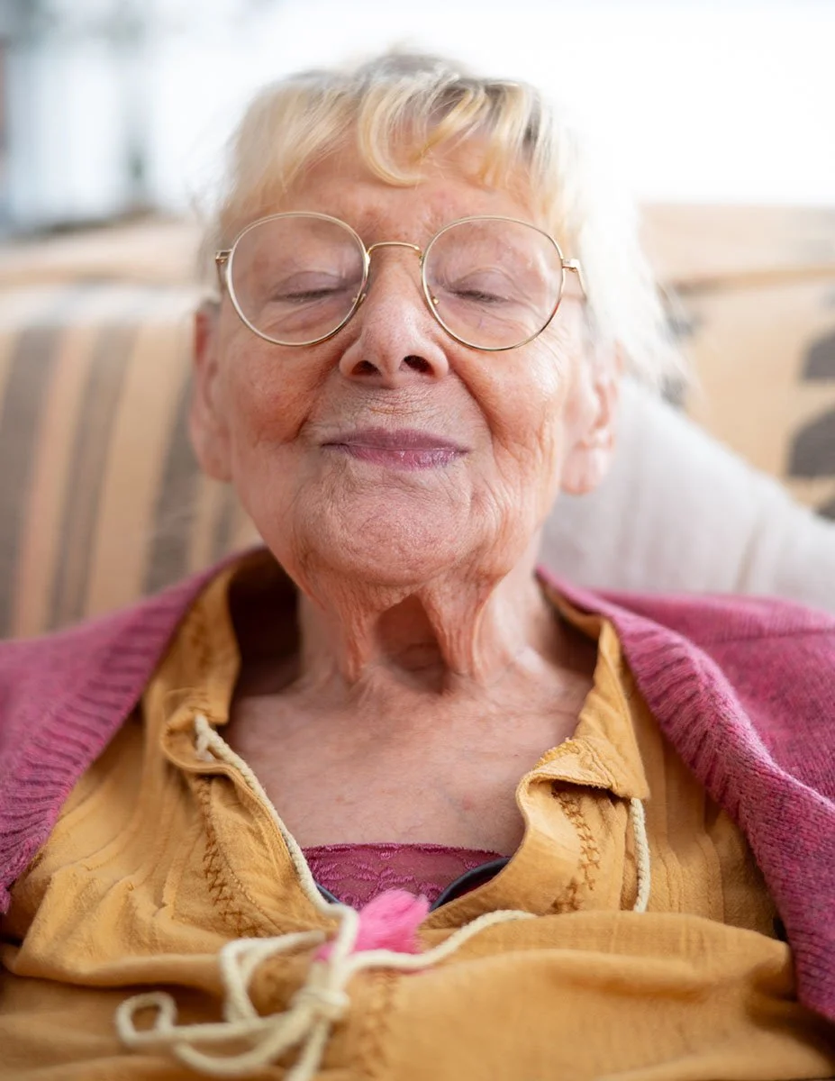 Ältere Frau mit geschlossenen Augen und Brille lächelt. Sie trägt eine gelbe Bluse, einen rosa Pullover und sitzt auf einem Sofa.