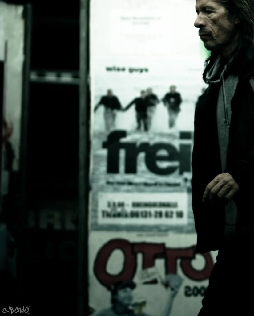 Ein Mann mit dunklen Haaren und Bart steht seitlich vor einer Wand mit Postern, im Hintergrund sind vier Personen zu sehen, die Händchen halten, das Bild ist unscharf.