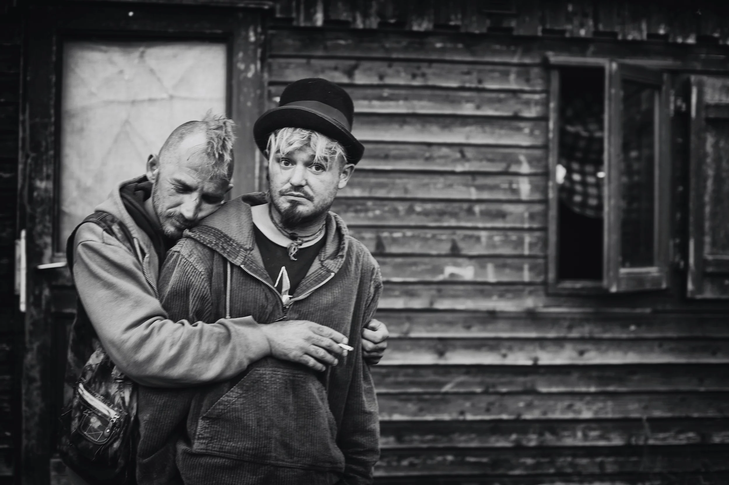 Zwei Männer stehen vor einem alten Holzhaus, einer der Männer trägt einen Hut und raucht, der andere lehnt mit dem Kopf auf dessen Schulter, beide wirken nachdenklich.