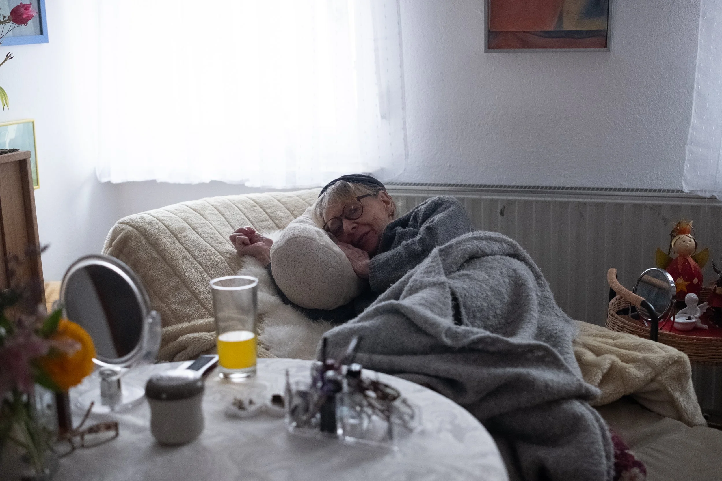 Ältere Frau liegt auf einem Sofa und schläft, eingehüllt in eine graue Decke, mit einem Kissen unter dem Kopf, in einem hell erleuchteten Raum.