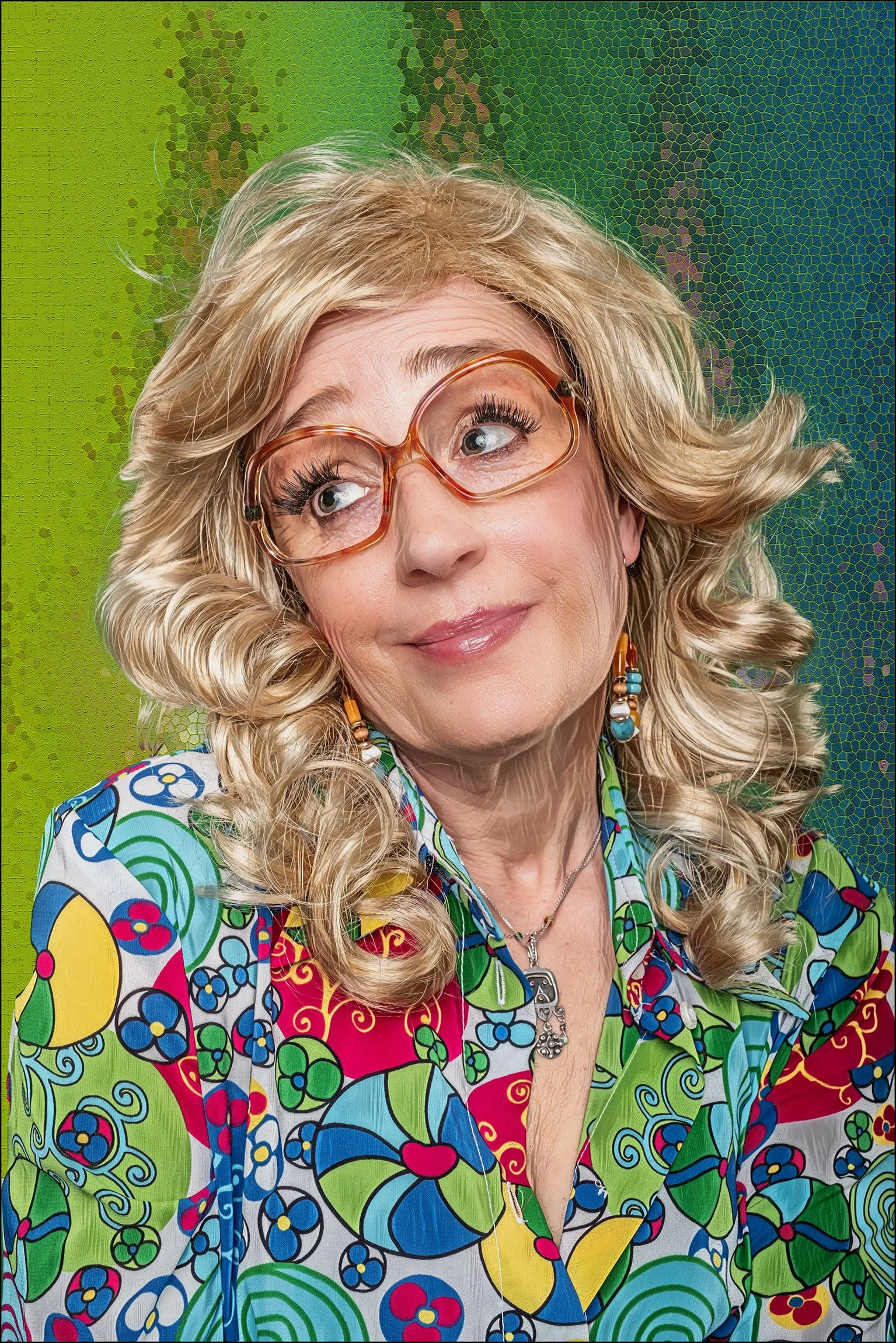 Dieses Bild zeigt eine Frau mit lockigem blondem Haar, die eine bunte Bluse mit Pflanzen- und Blumenmuster trägt. Sie trägt große, transparente Brillengläser, bunte Ohrringe und ein Halsband mit Anhängern, während sie nach oben schaut mit einem leich