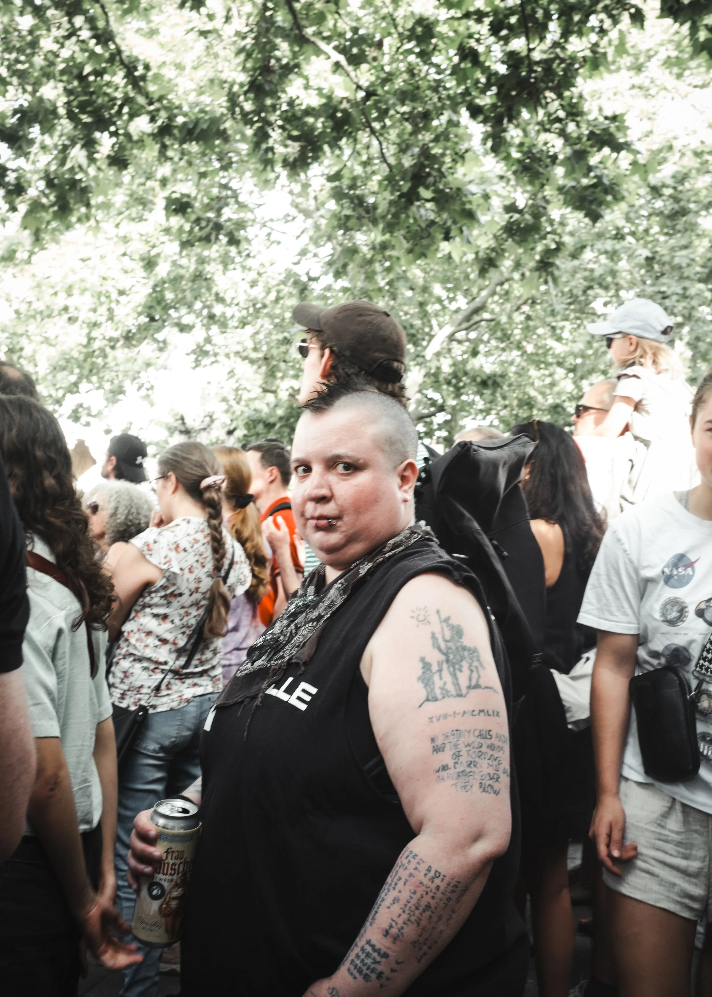 Person mit kurzen Haaren, Tattoos auf Armen, schwarze Kleidung, umgeben von Menschen bei einer Demonstration gegen rechts unter Bäumen.