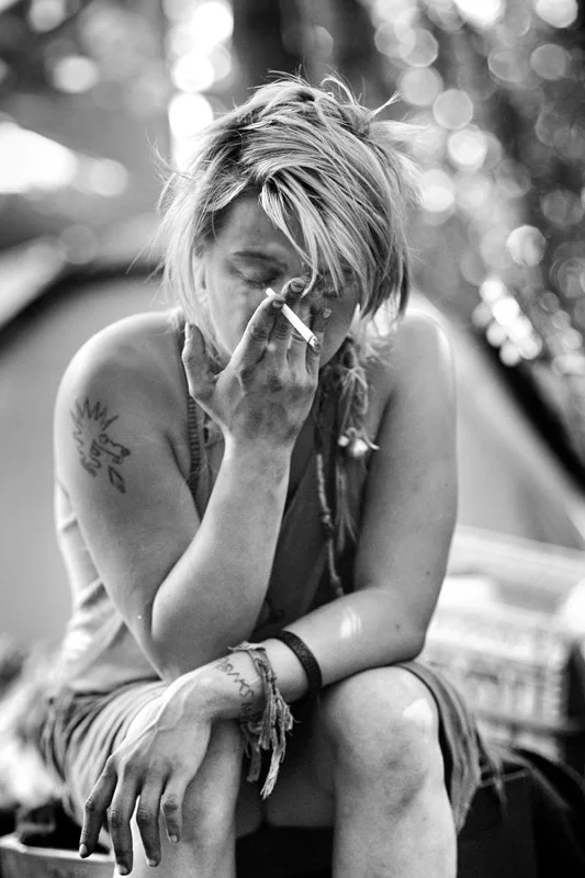 Schwarz-weiß Foto einer Frau mit kurzen, zerzausten Haaren, die an einem Zelt im Freien sitzt und eine Zigarette raucht.