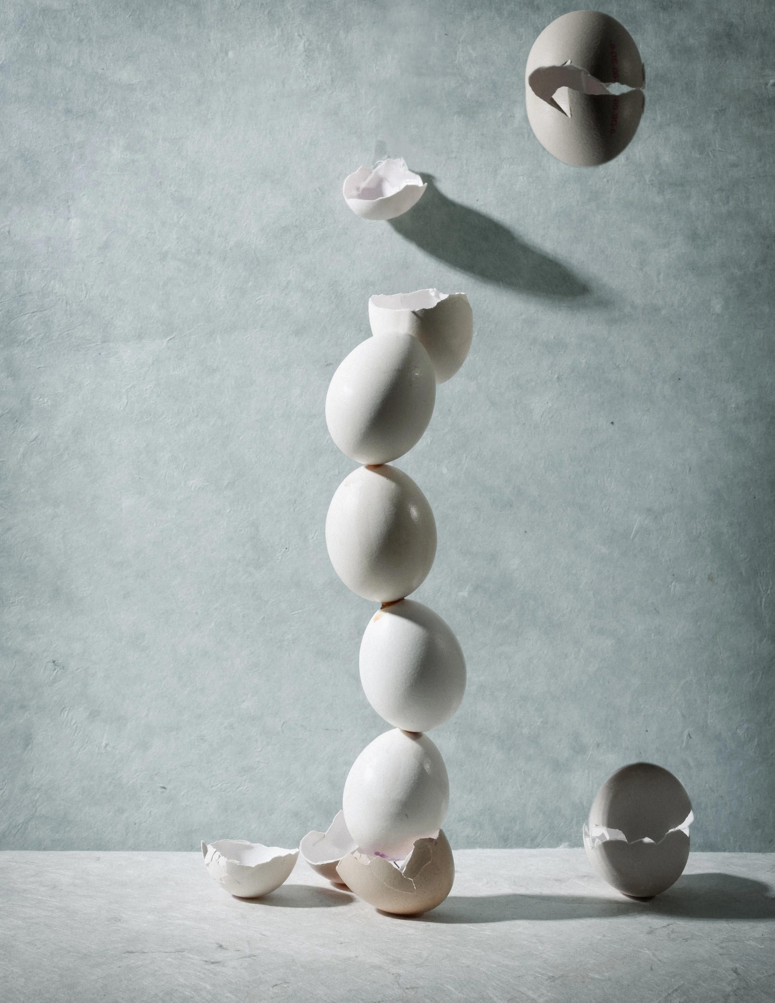 Fünf weiße Eier, die in einer vertikalen Linie gestapelt sind, mit einigen zerbrochenen Eierschalen um sie herum, und ein einzelnes halbiertes Ei in der Luft.