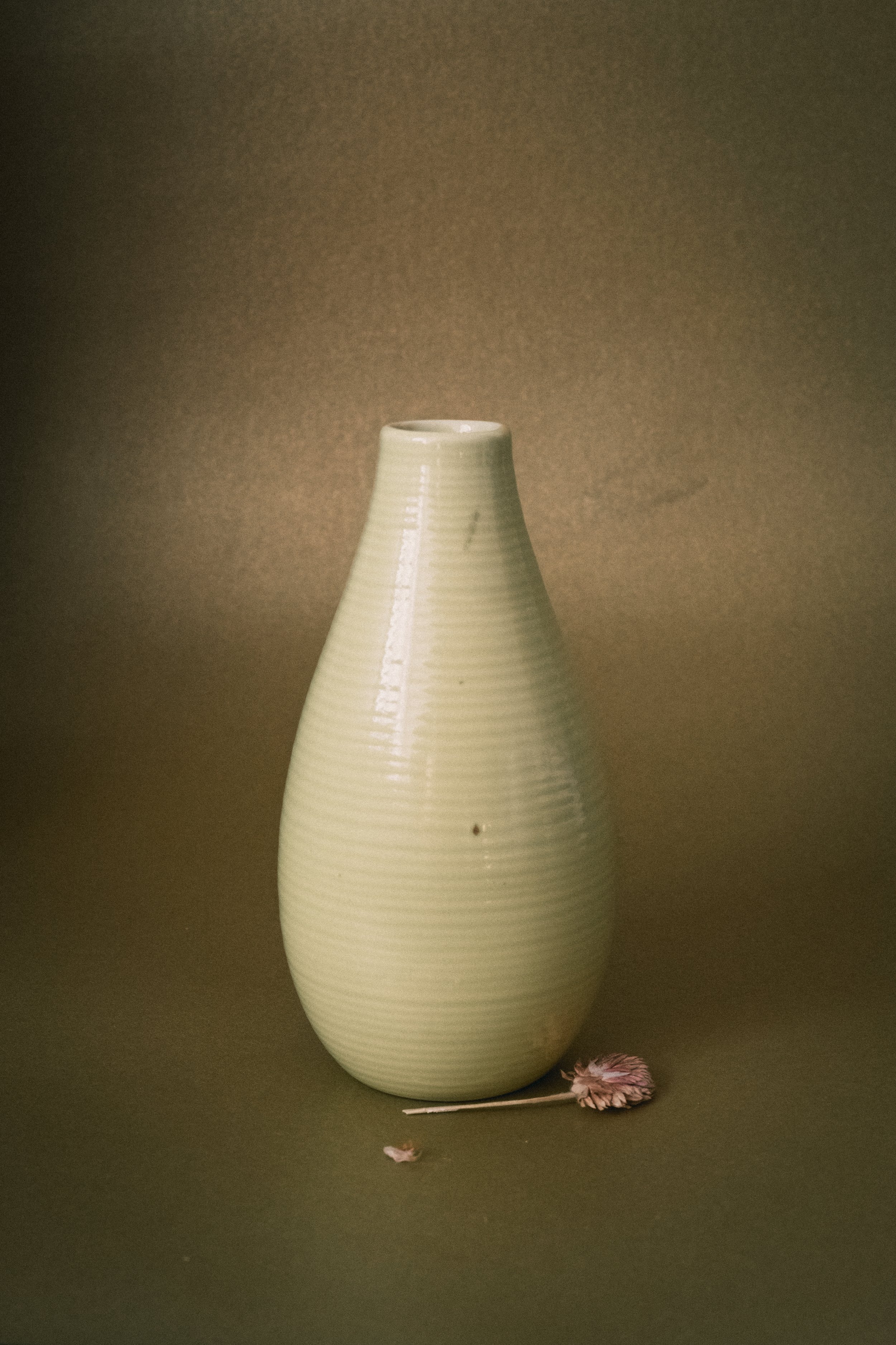 Eine hellgrüne Vase auf einem braunen Hintergrund mit getrockneten Blumen nebendran.