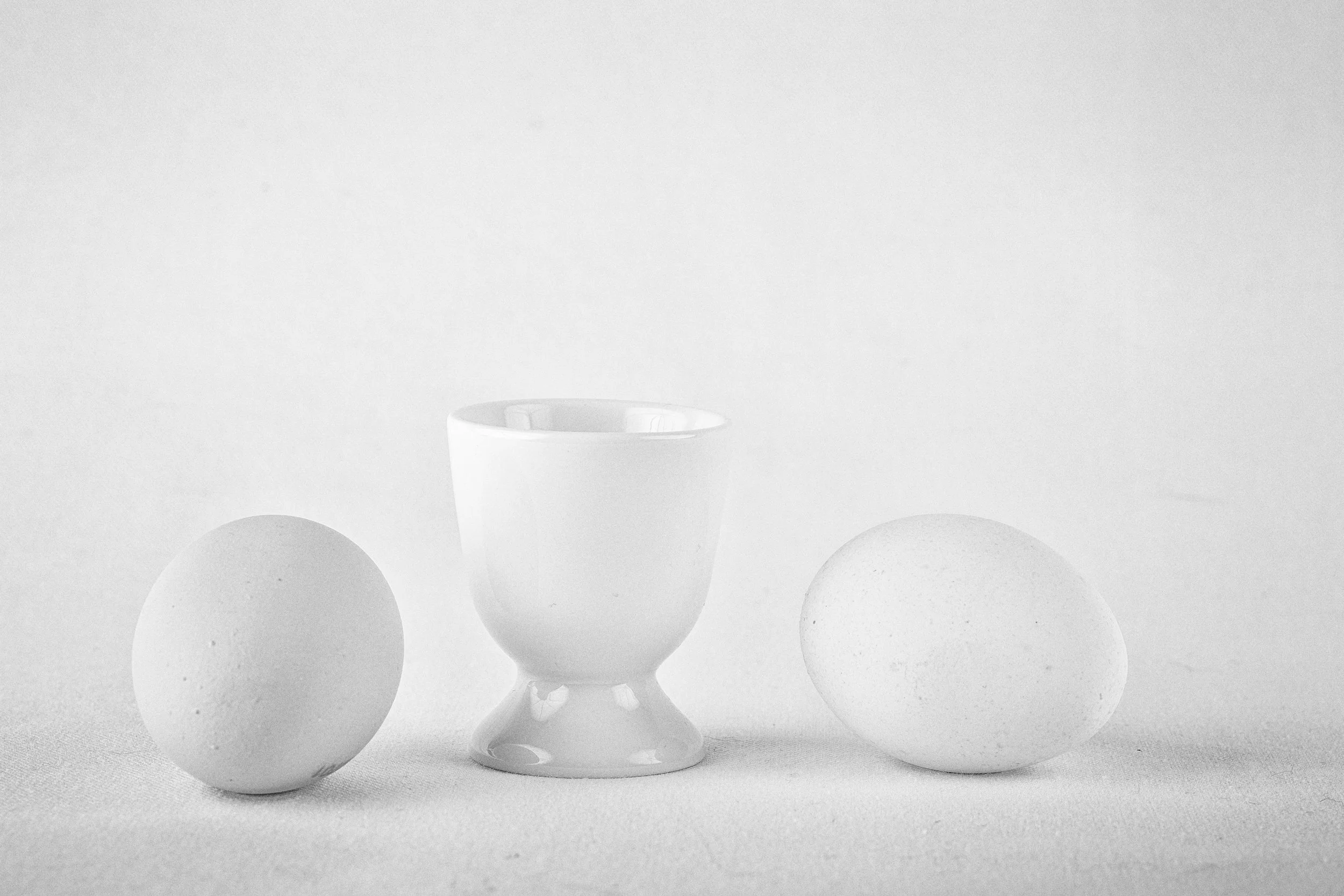 Zwei weiße Eier liegen auf einem weißen Tuch, zwischen ihnen steht ein weiße Eierbecher.