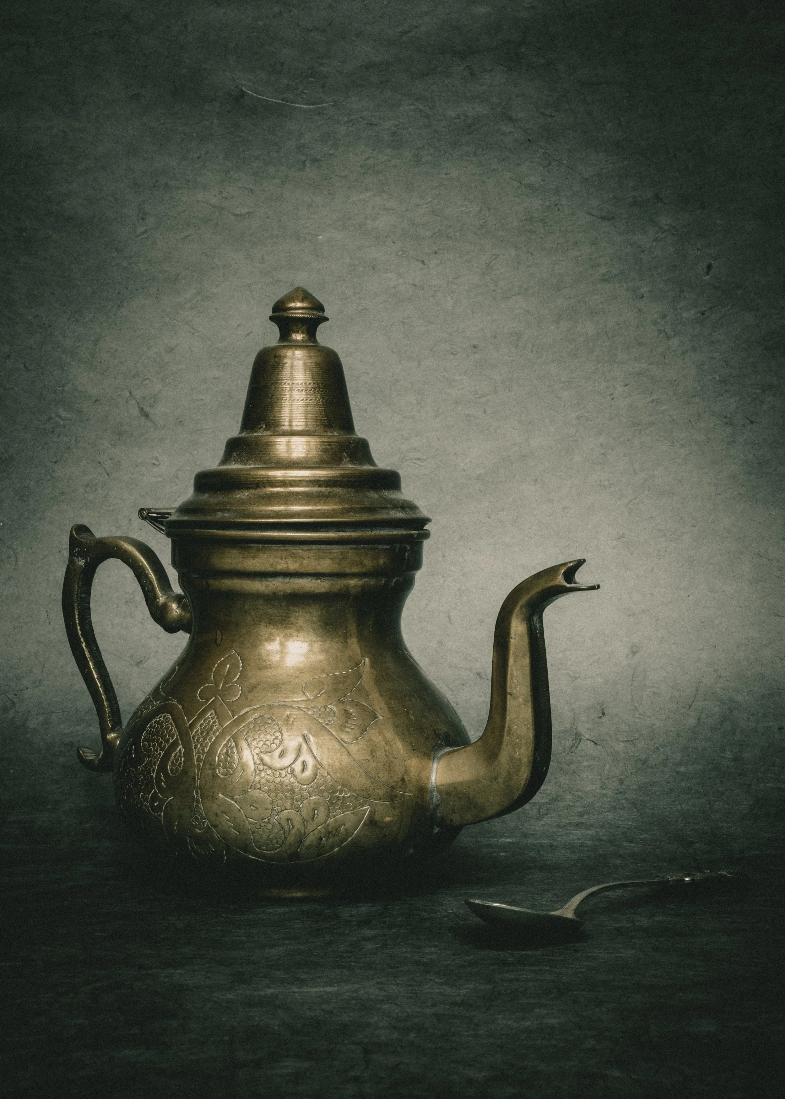 Ein antiker, goldener Teekessel mit floral bemusterter Oberfläche, daneben ein silberner Löffel auf dunklem Untergrund.