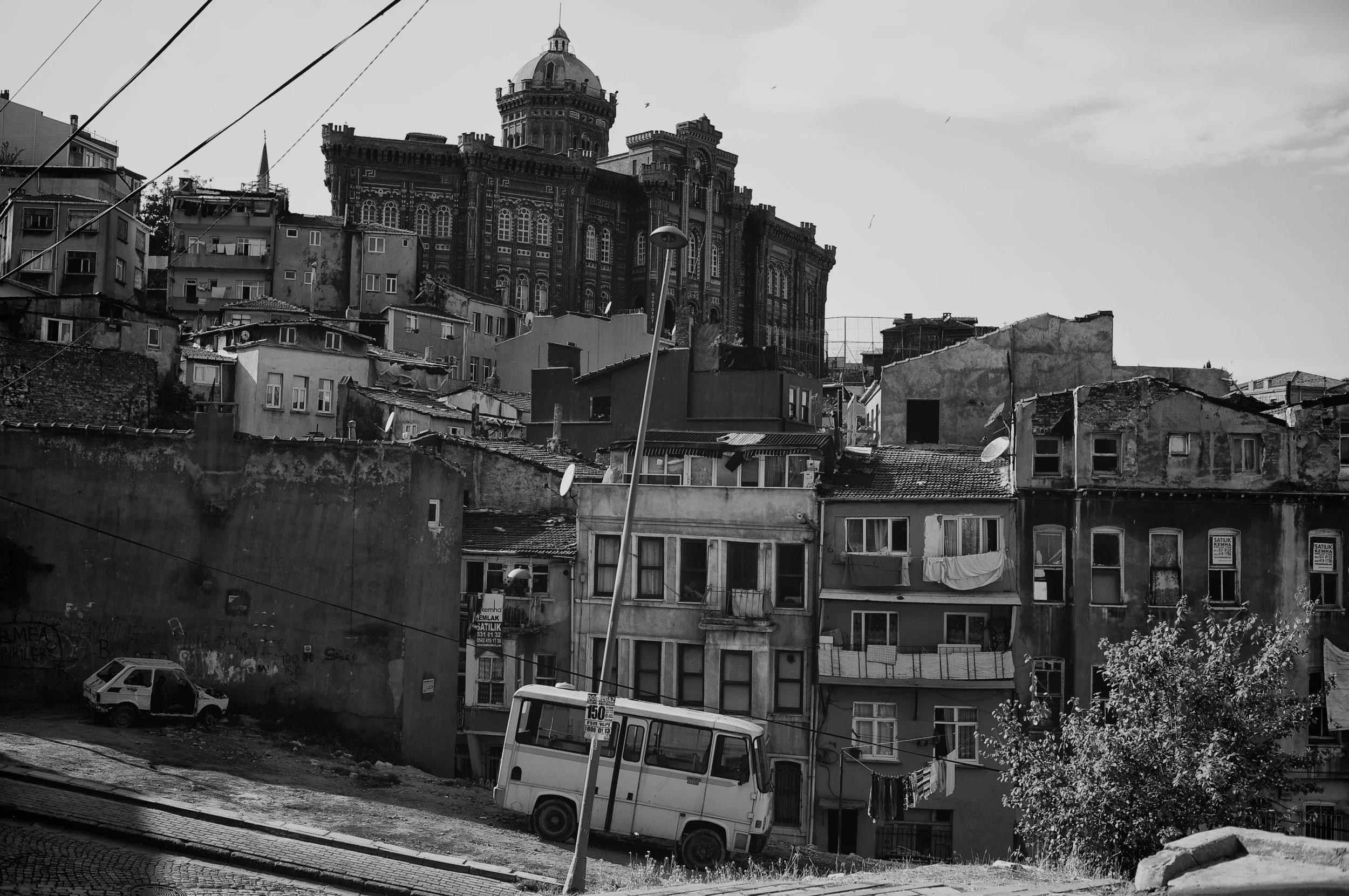 Eine Stadt mit alten und beschädigten Wohnhäusern im Vordergrund und einem großen, historischen Gebäude auf einem Hügel im Hintergrund, alles in schwarz-weiß.