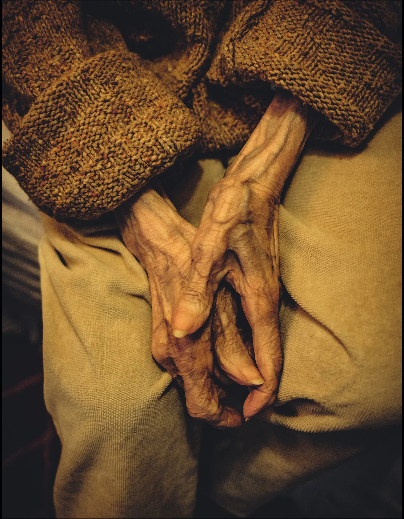 Nahaufnahme der hinkenden Hand eines älteren Menschen, der einen braunen, gestrickten Pullover trägt und beige Hosen.