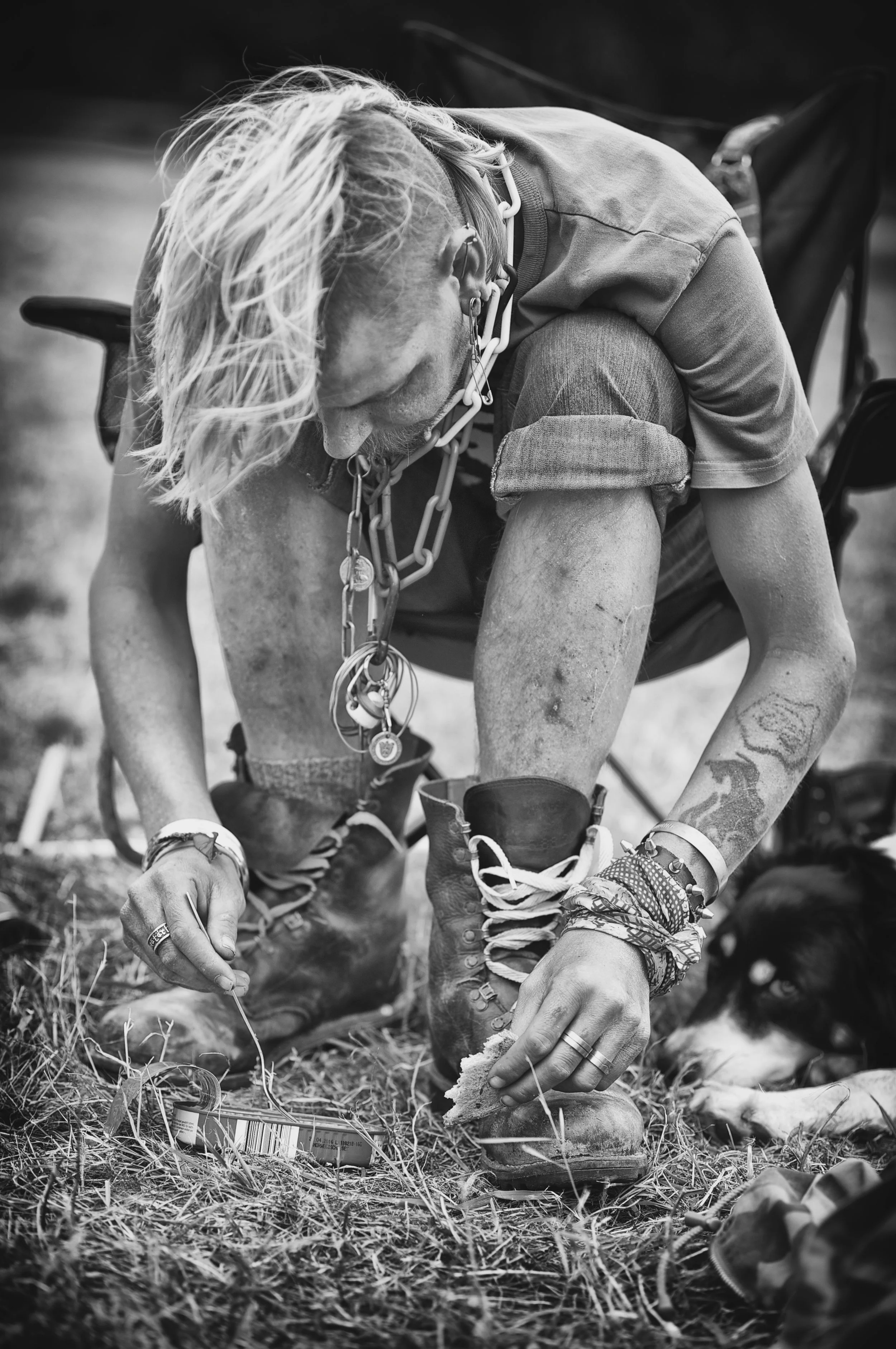 Schwarz-weiß-Fotografie eines Mannes mit Tattoos und langen Haaren, der auf dem Boden sitzt und eine Schnur an einem ID-Tag befestigt, während ein Hund neben ihm liegt.