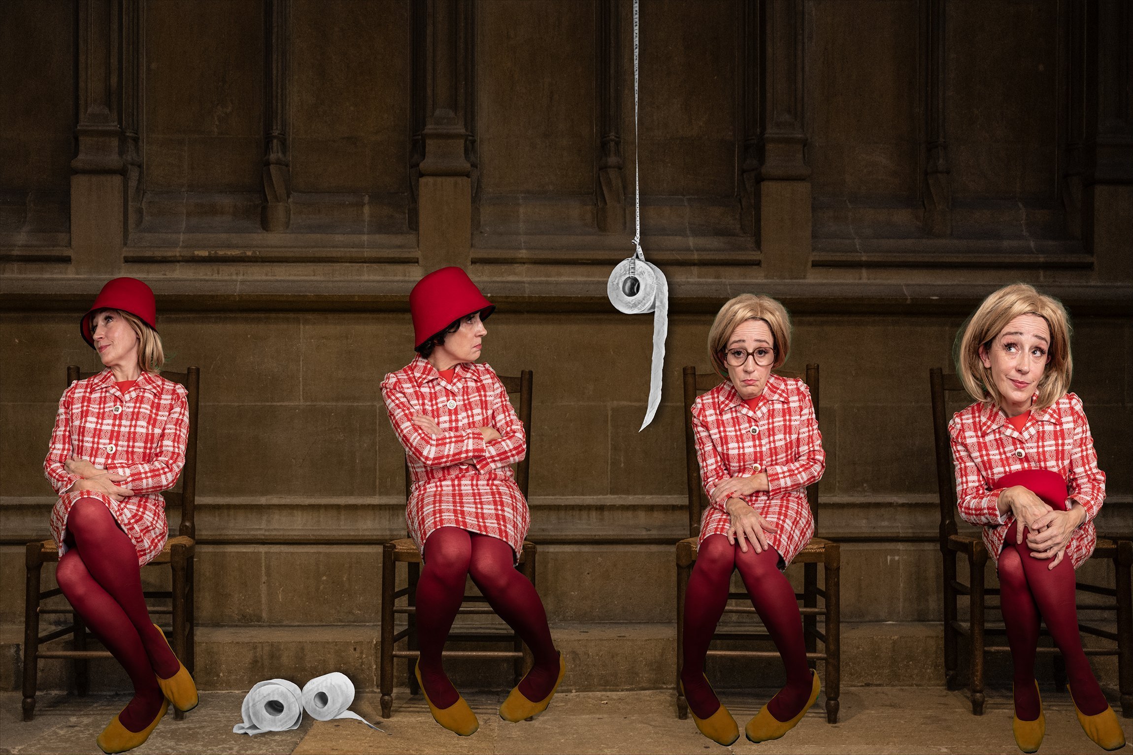 Vier Frauen sitzen auf Stühlen in einer Reihe in einem Raum mit Holzwänden. Sie tragen rote Kittelschürzen, rote Strumpfhosen, gelbe Schuhe, und unterschiedliche rote Hüte. In der Mitte hängt eine Rolle Toilettenpapier an einem langen Faden von der D