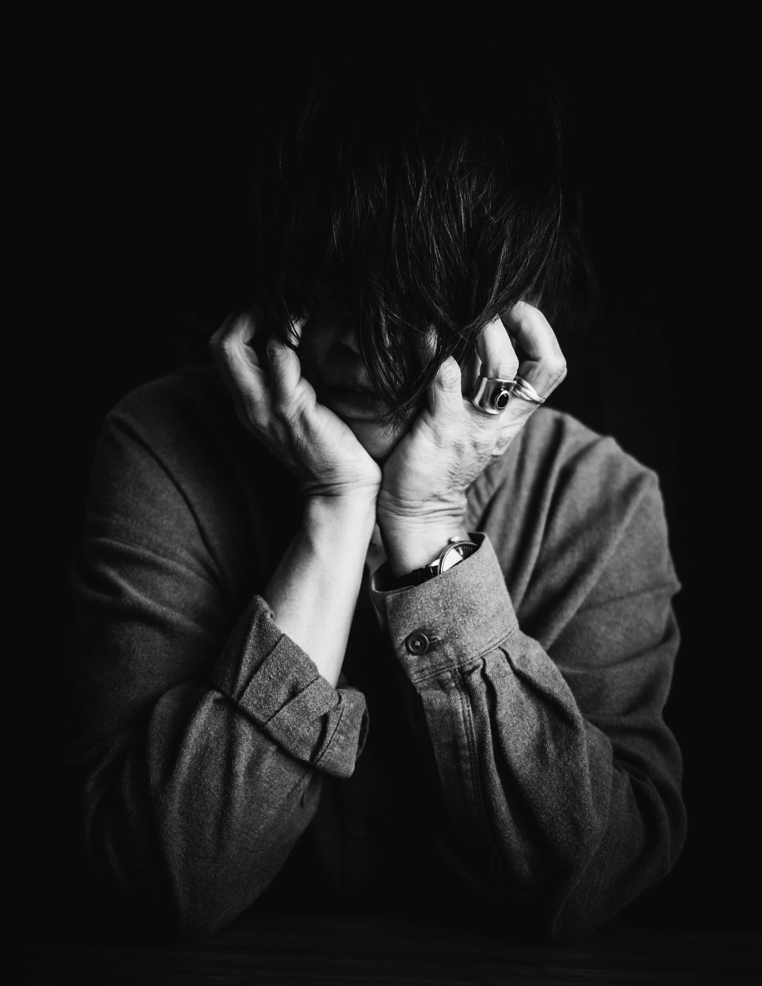Schwarz-Weiß-Foto einer Person mit verschränkten Händen vor dem Gesicht, Haare verdecken die Mimik, dunkler Hintergrund.