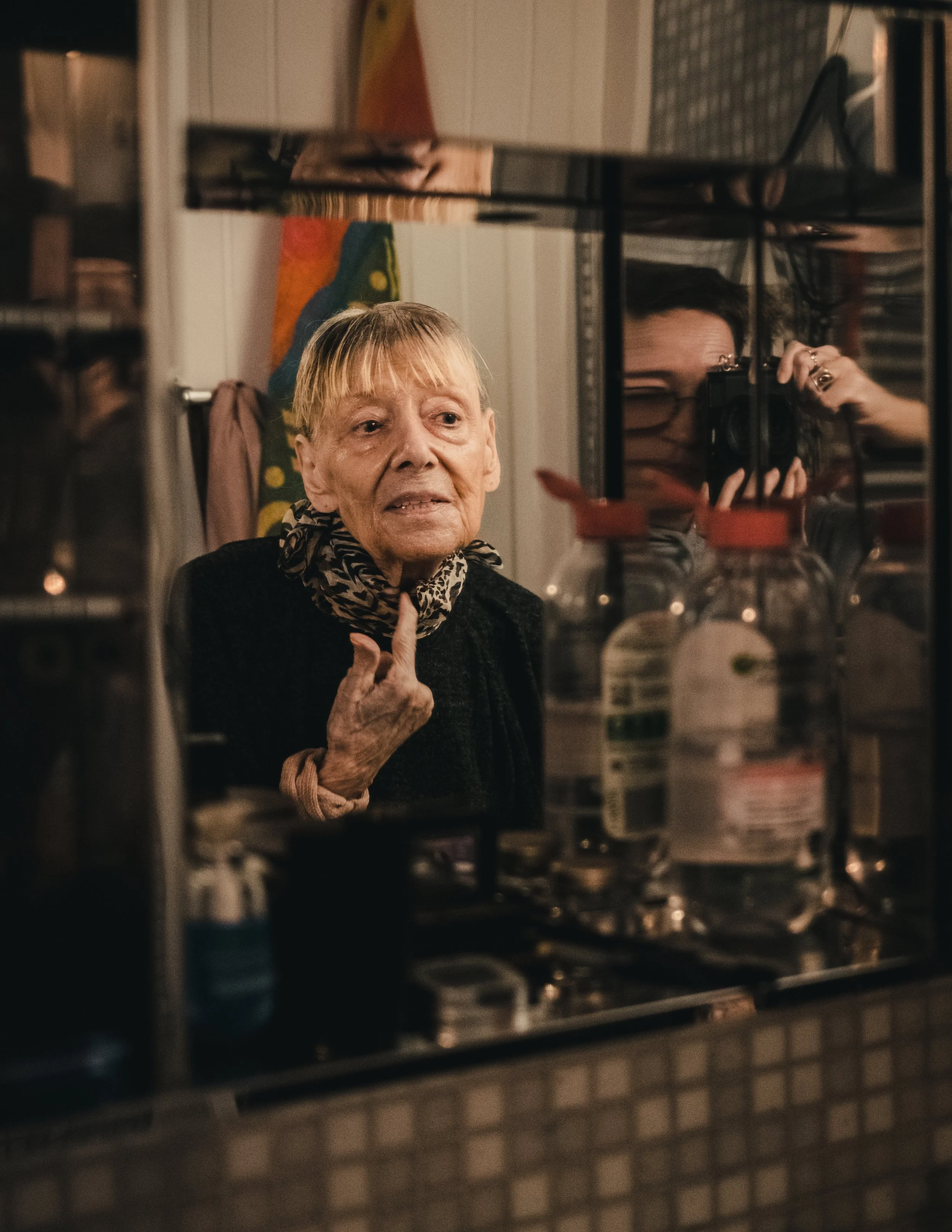 Ältere Frau schaut in einen Spiegel und zeigt mit dem Finger auf ihr Kinn, während eine Person im Hintergrund Fotos macht.