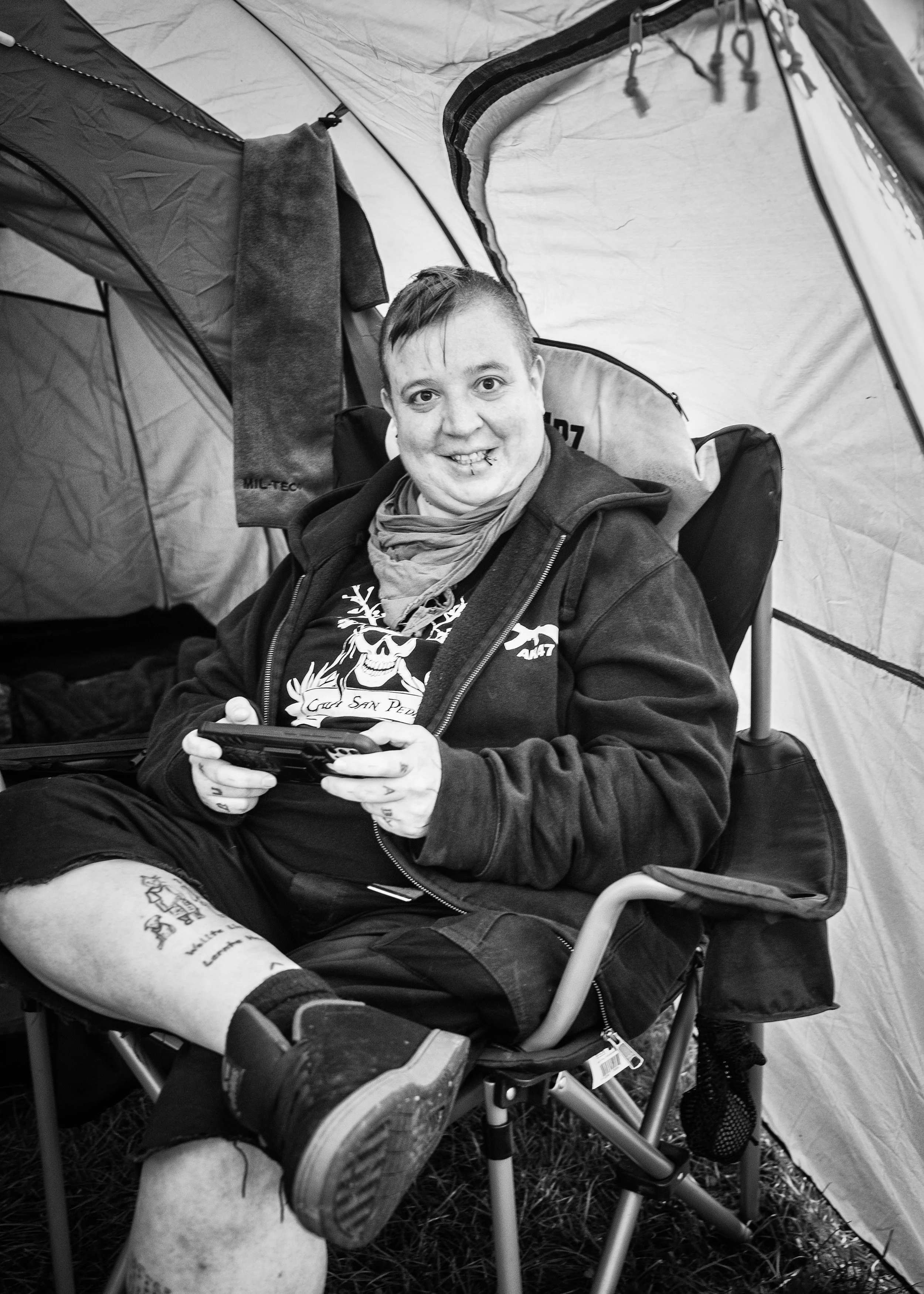 Junger Mann sitzt in einem Campingstuhl in einem Zelt, lächelt und hält ein Smartphone in den Händen.