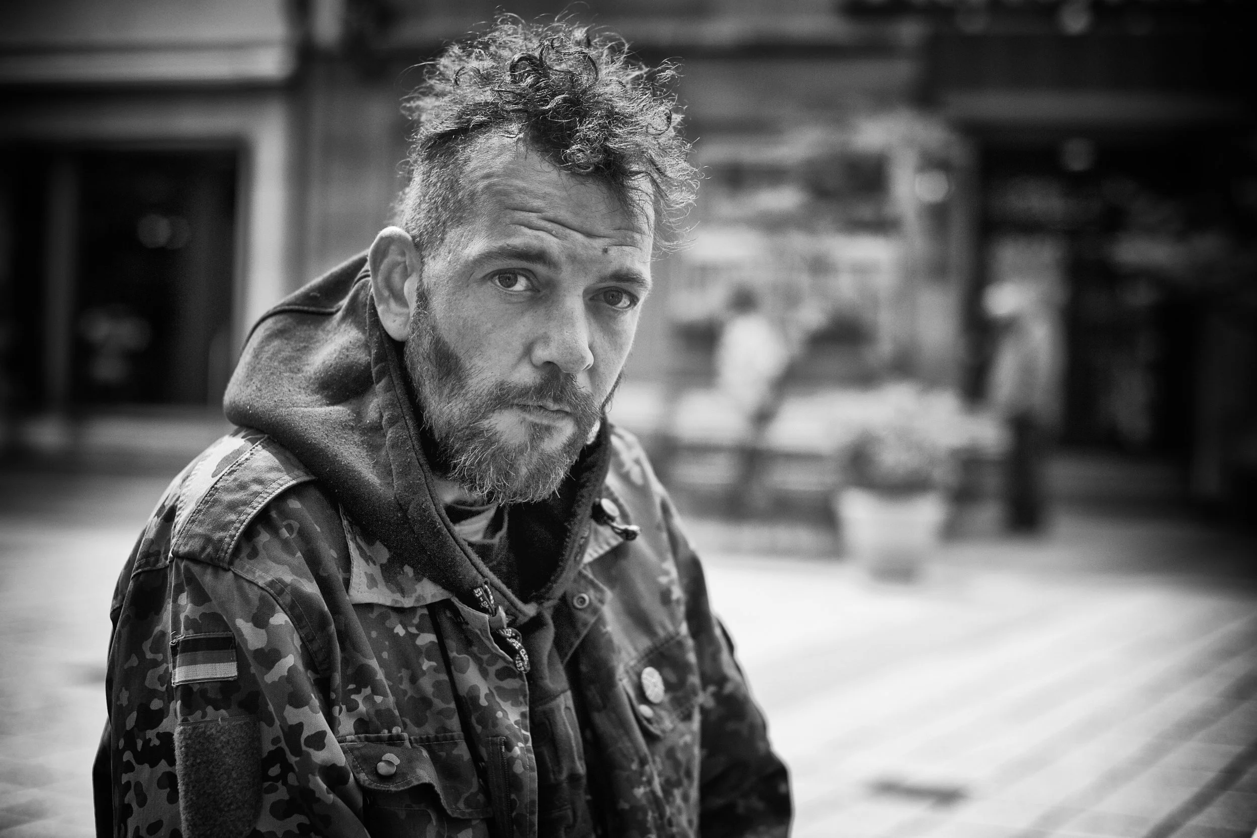 Schwarz-Weiß-Foto eines Mannes mit vollem Bart und lockigem Haar, trägt eine Camouflage-Jacke, blickt ernst in die Kamera, im Hintergrund eine Person und ein Blumenkübel, unscharf.