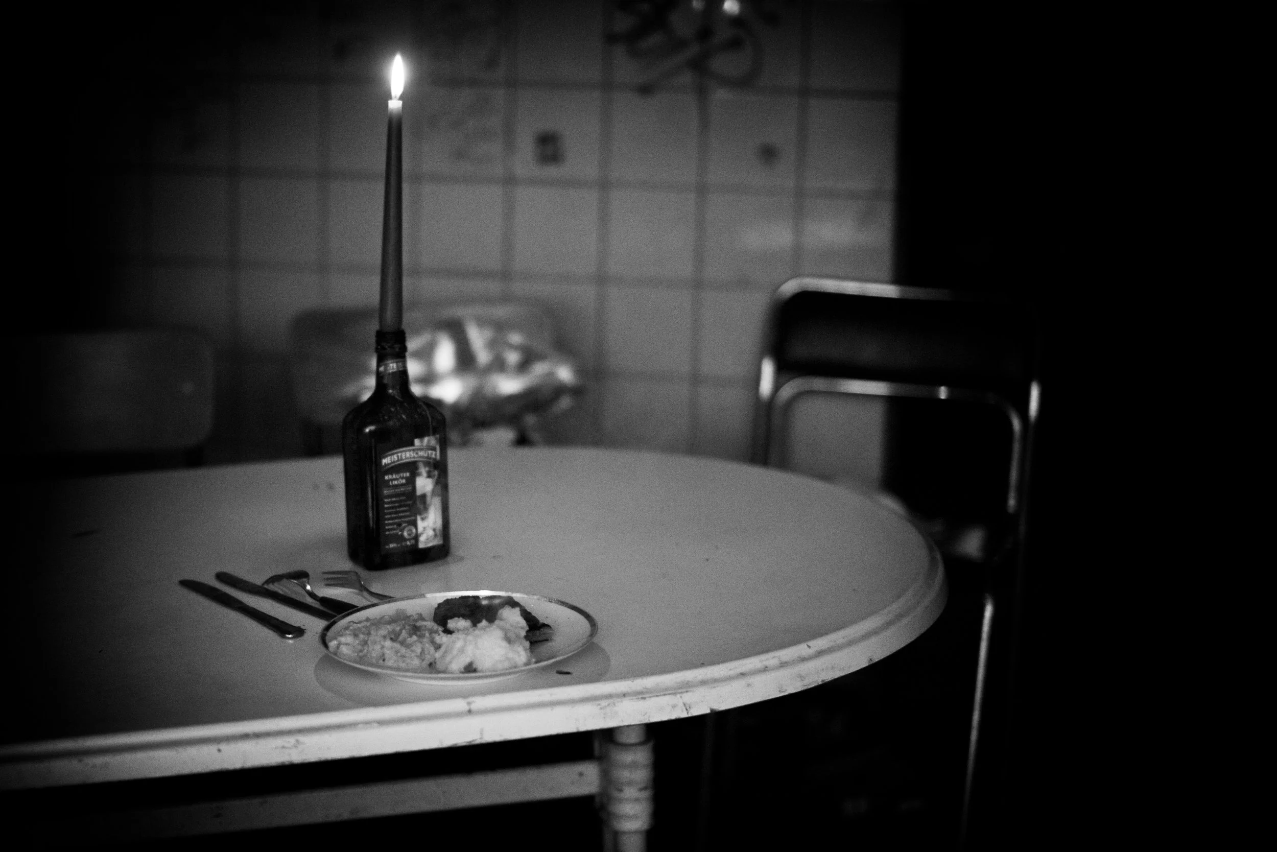 Ein runder Esstisch mit einer leeren Mönchsglas-Flasche, die eine brennende Kerze hält, und einem Teller mit Essen. Im Hintergrund sind kahle Wände mit Fliesen und ein Stuhl.