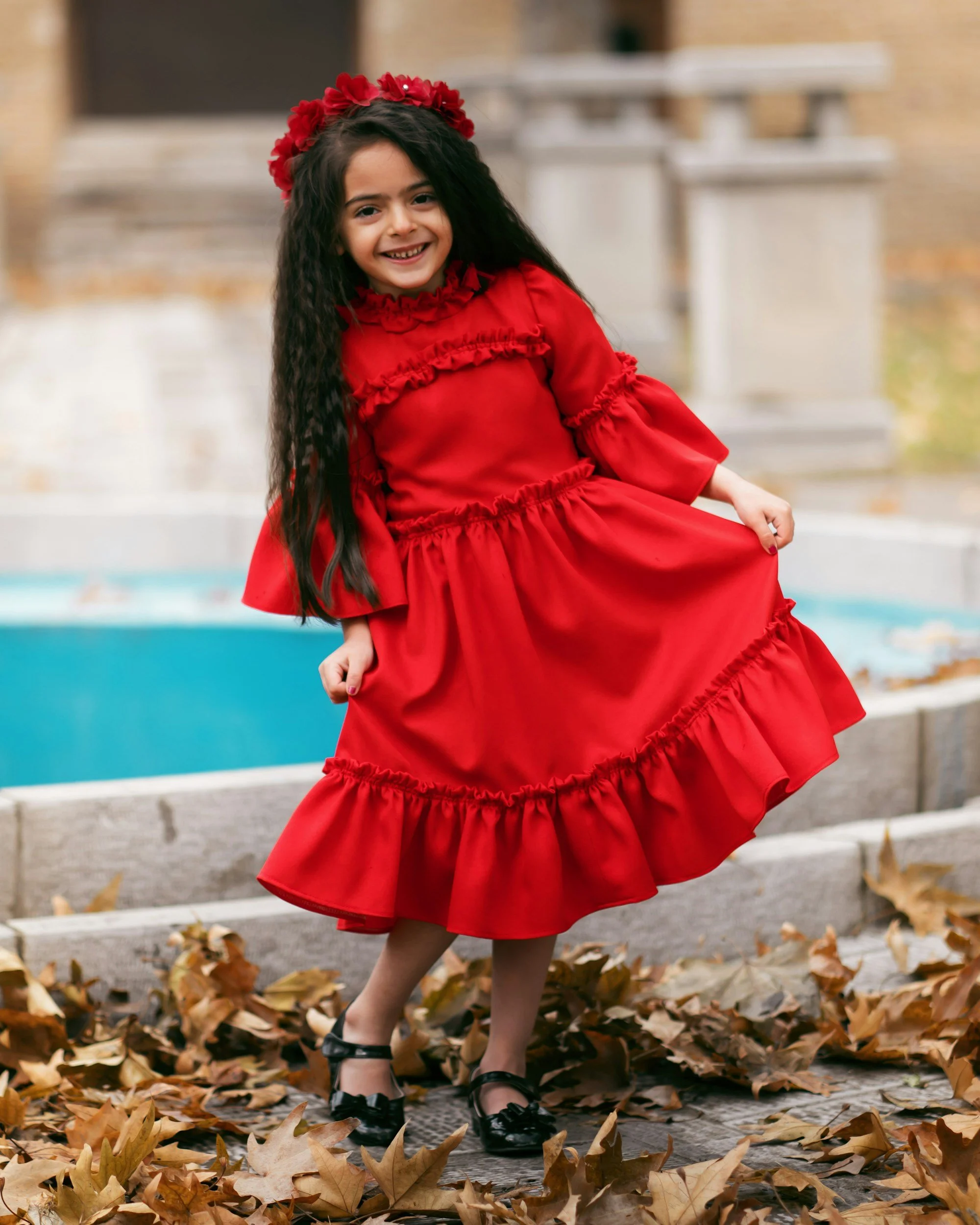 A young girl with long dark hair, smiling, wearing a red dress with ruffles and a matching floral headband, standing outdoors amidst fallen leaves near a fountain.
