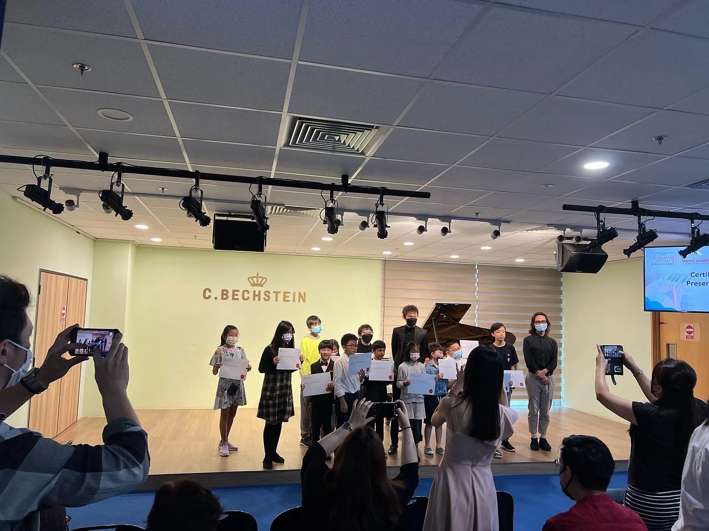 Children and adults stand on a stage holding certificates during an award ceremony or recital in a music hall. A grand piano and the name 'C. Bechstein' are visible in the background, along with audience members taking photos.