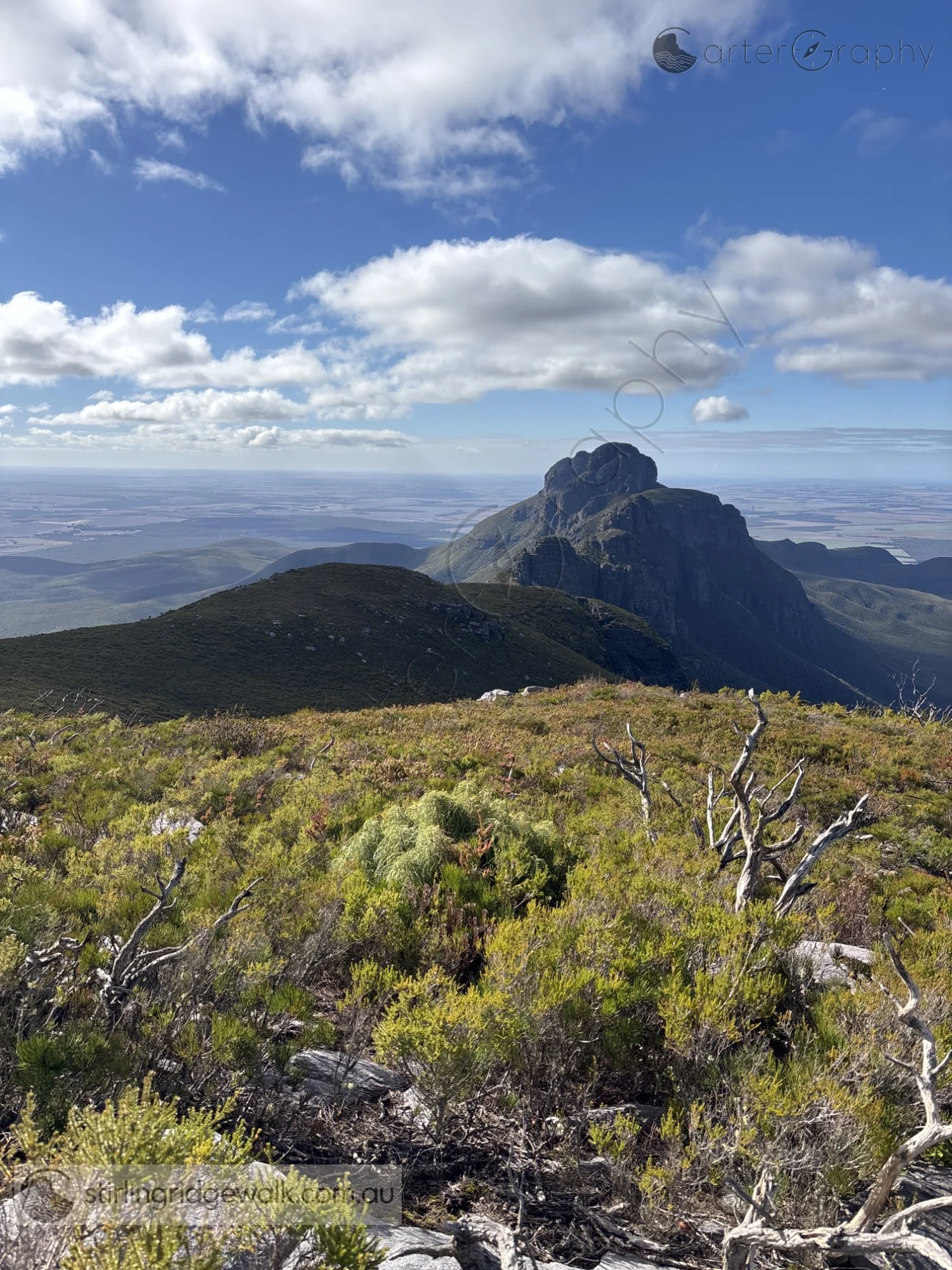 Looking east back along the Stirling Ridge Walk from the approach to Isongerup Peak. A broad rounded ridge fills the middle ground, leading towards the rugged Arrow formations beyond, with the farmland and plains stretching away to the north.