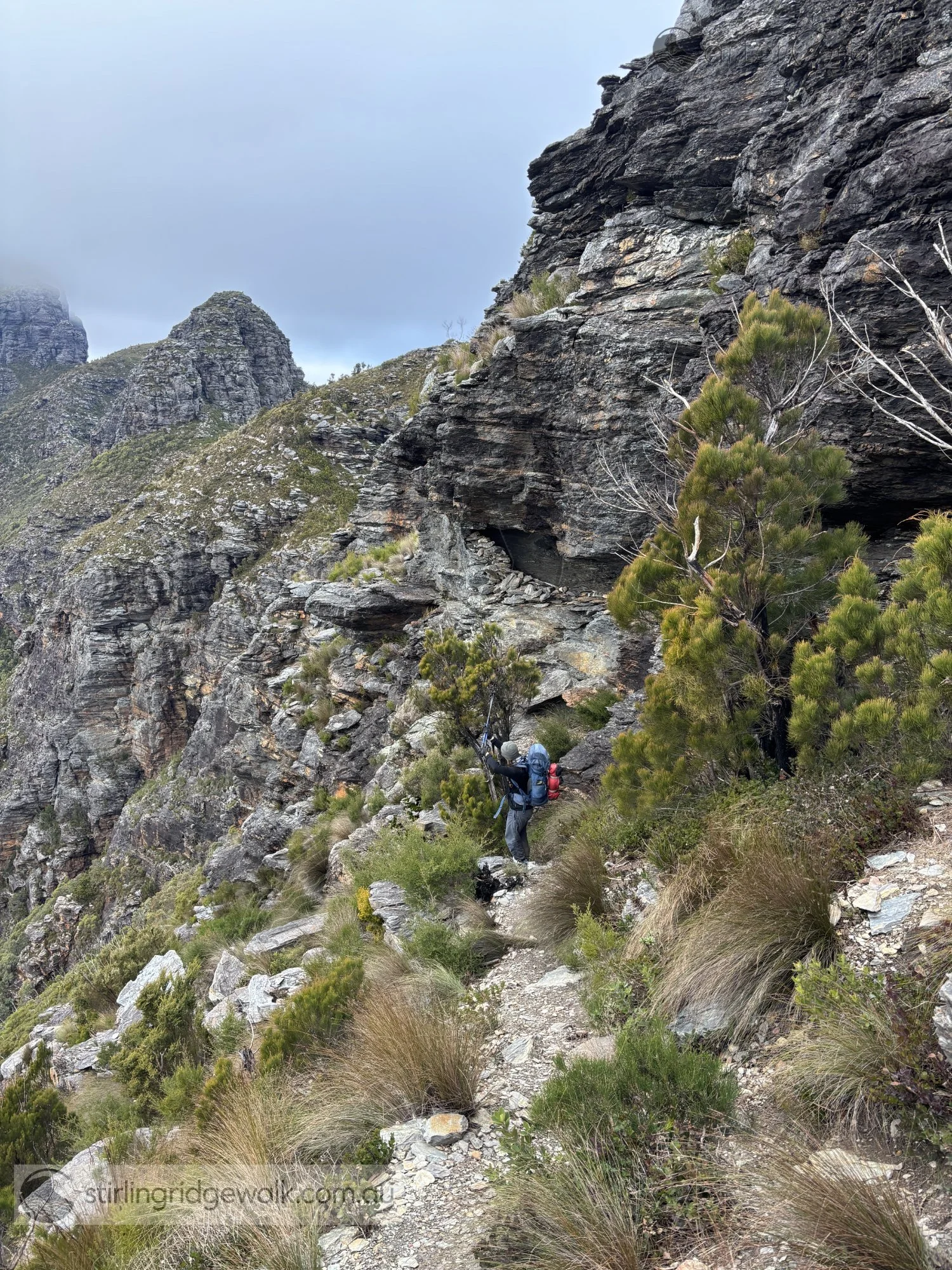 A walker passing First Arrow Cave after descending from the summit of 1st Arrow, with 2nd Arrow and 3rd Arrow visible beyond and Pyungoorup Peak partly obscured by cloud in the upper left background.