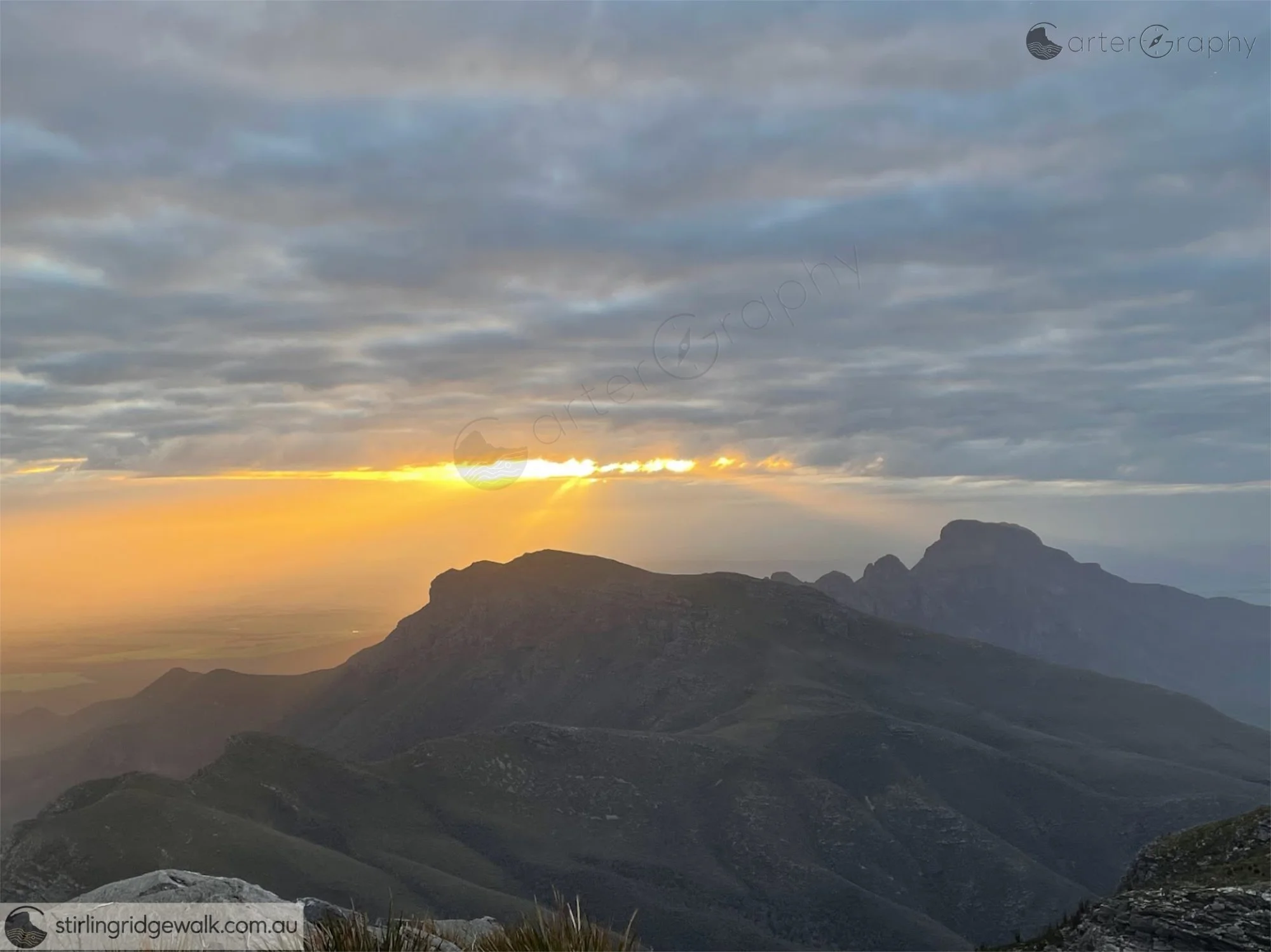 Looking ENE from East Peak as the setting sun breaks beneath a heavy layer of cloud, casting warm light across the plains. The successive peaks and rocky features of the Stirling Ridge Walk recede into silhouette towards Pyungoorup Peak in the far di
