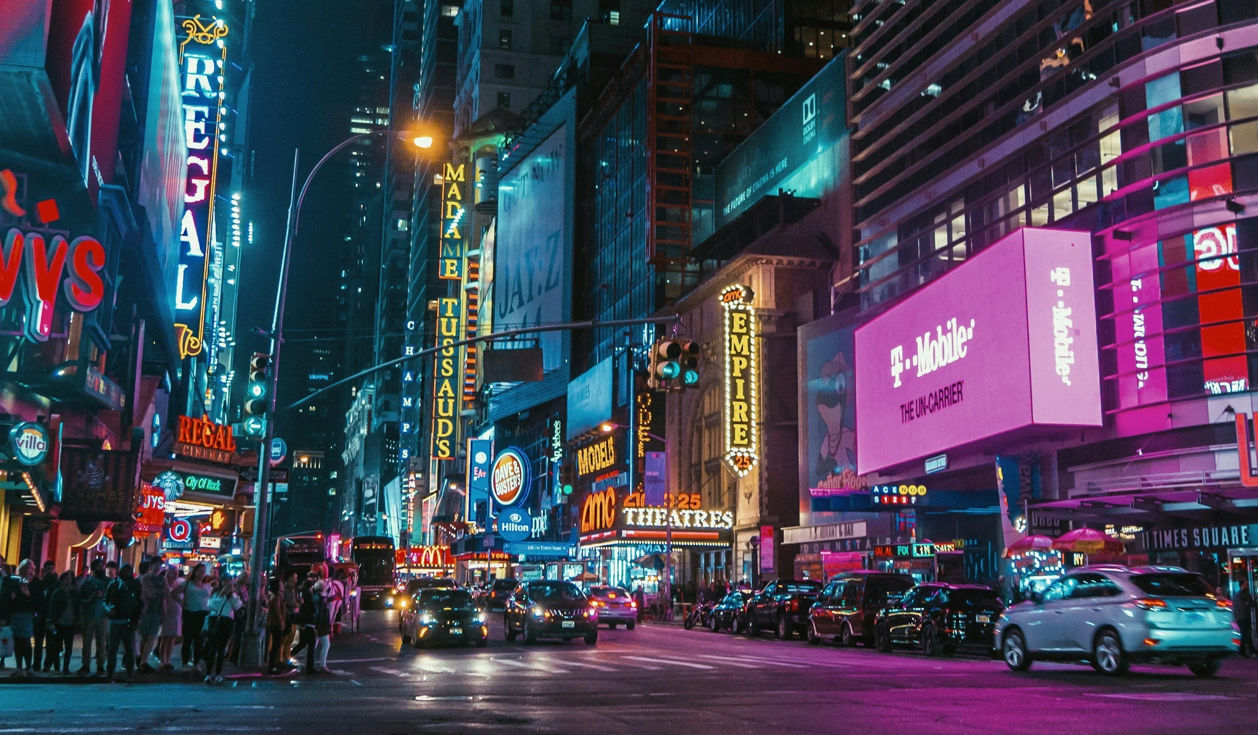 Brightly lit Times Square at night with neon signs, billboards, and a crowd of pedestrians and cars.