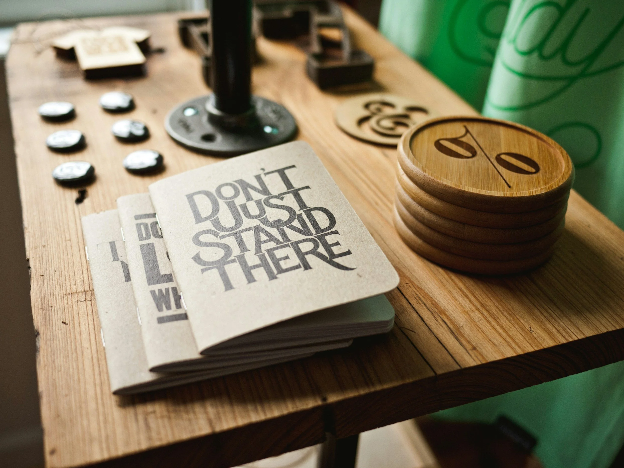A wooden table with game pieces, a stack of wooden Coasters with a zero and divided by symbol on the top, a small open notepad with the phrase ''Don't Just Stand There'' printed on the page, and a green drink container in the background.