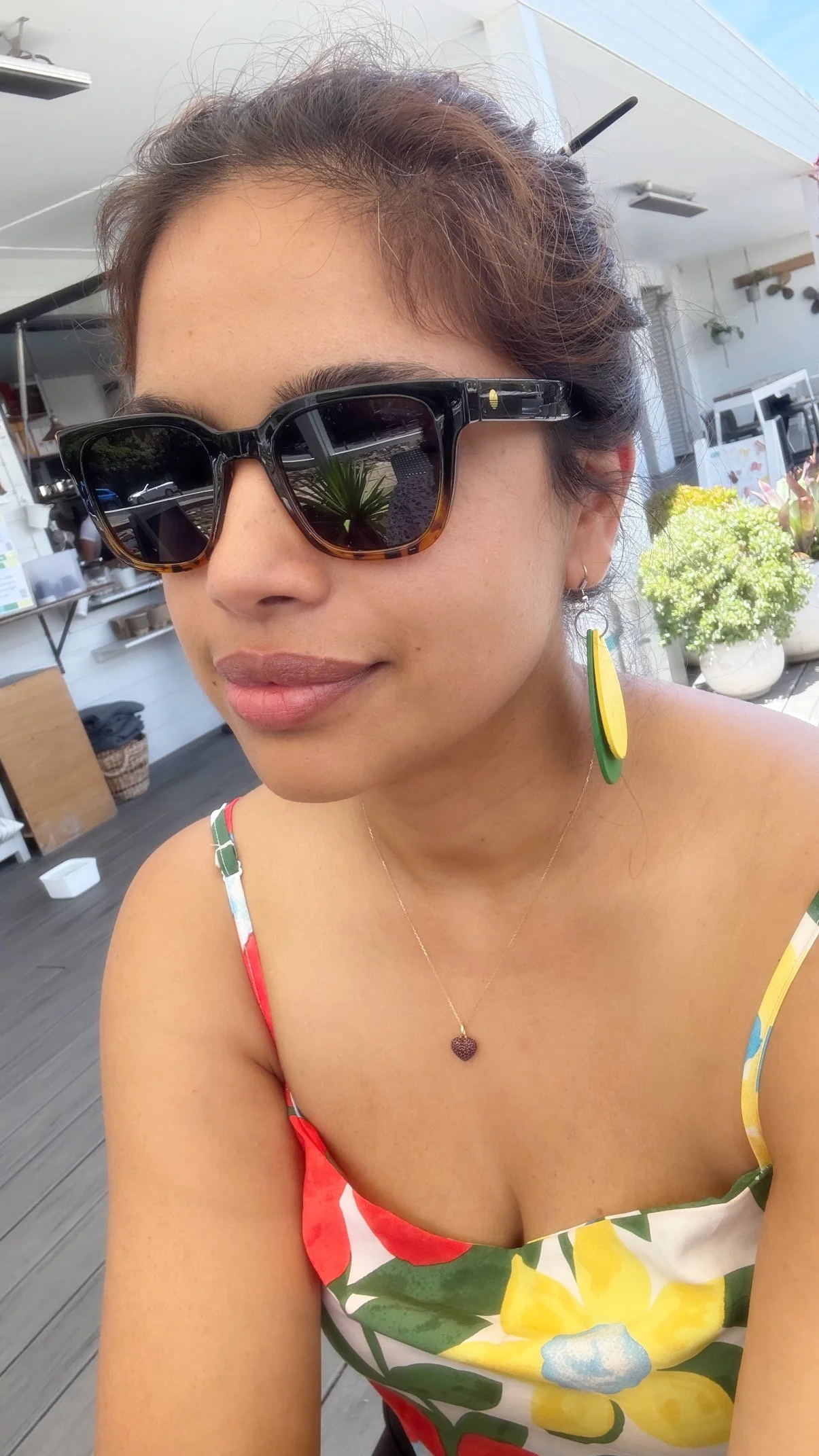 A woman with sunglasses, a colorful floral dress, and leaf-shaped earrings, sitting outdoors in a bright, sunny environment with plants and a white building in the background.