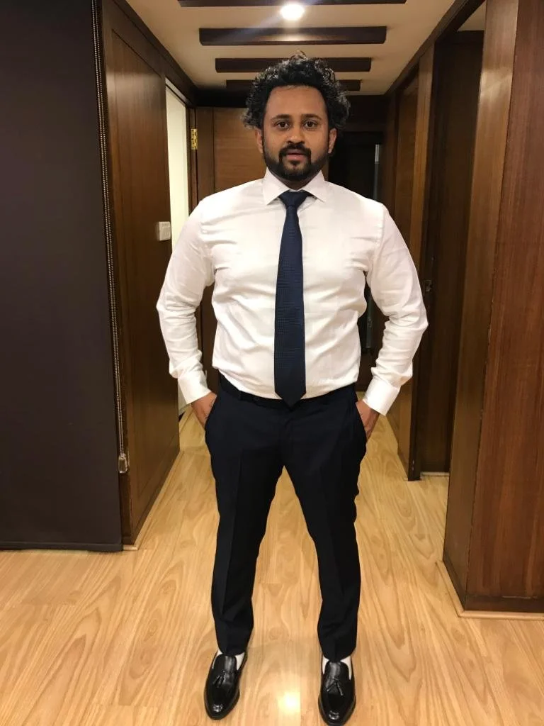 A man with curly hair and a beard wearing a white dress shirt, navy tie, and navy dress pants, standing in a wood-paneled hallway with his hands in his pockets.