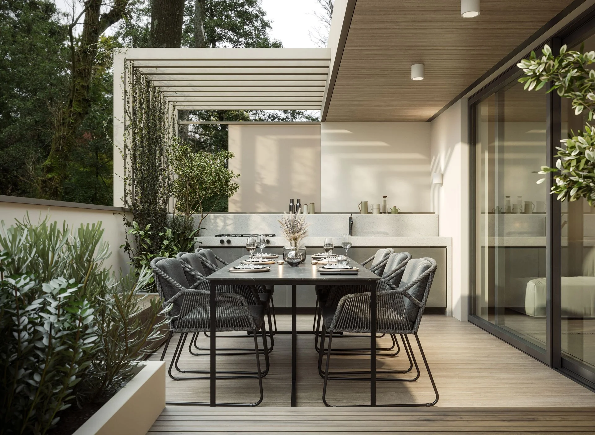 Modern outdoor dining area with a black dining table, gray chairs, glassware, and a vase of dried flowers, situated on a wooden deck with sliding glass doors leading inside, surrounded by greenery and a partial white pergola overhead.