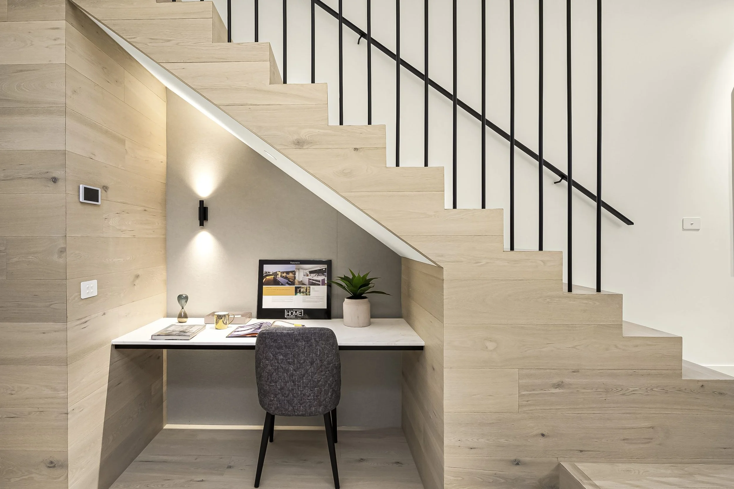 A modern workspace under a staircase with a white desk, gray chair, potted plant, computer monitor, and minimal decor.