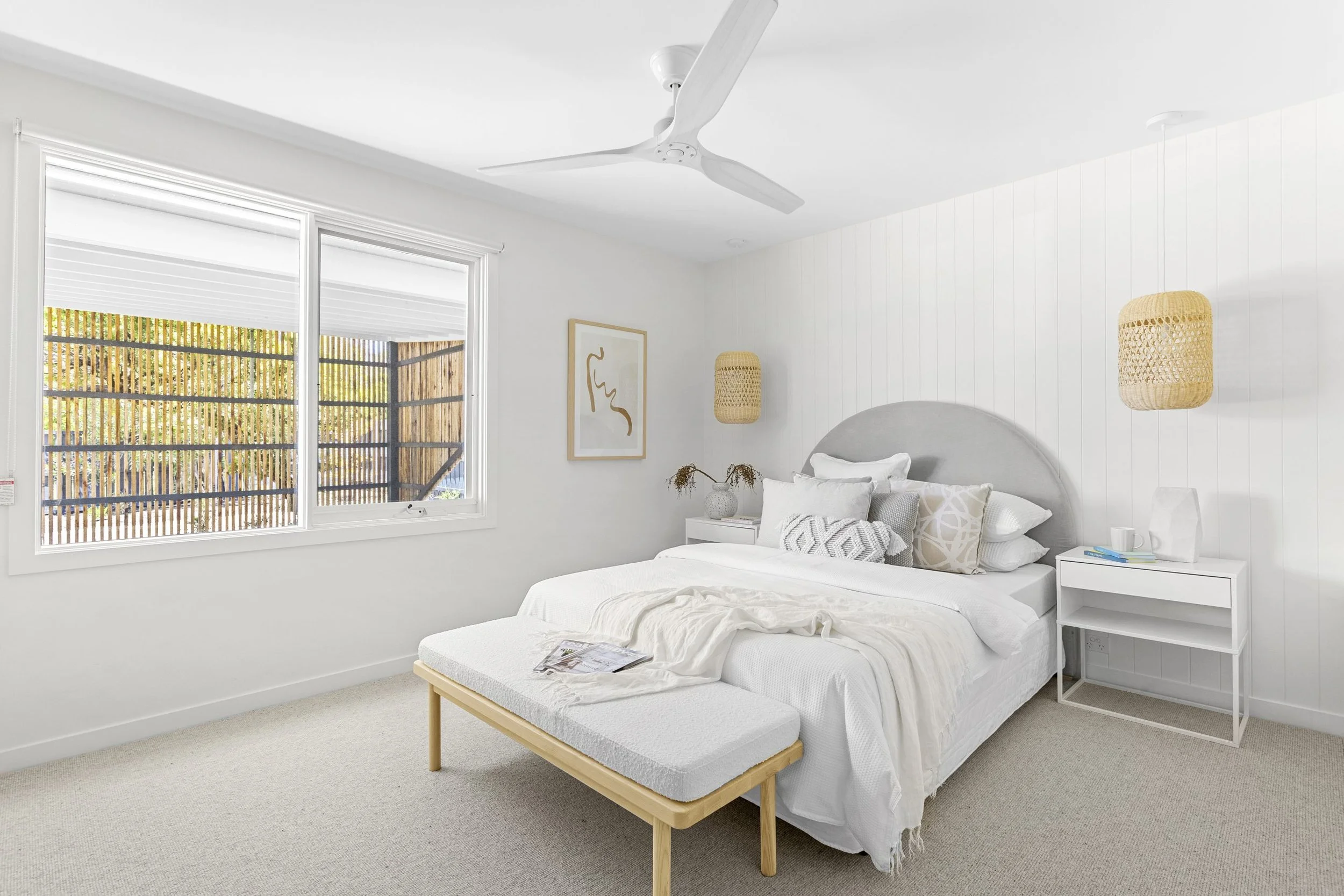 A minimalist bedroom with white walls, beige carpet, a bed with white linens and decorative pillows, a small white nightstand with items, wall-mounted woven lamps, a framed abstract art, a window with blinds, a ceiling fan, and a beige bench at foot of bed.