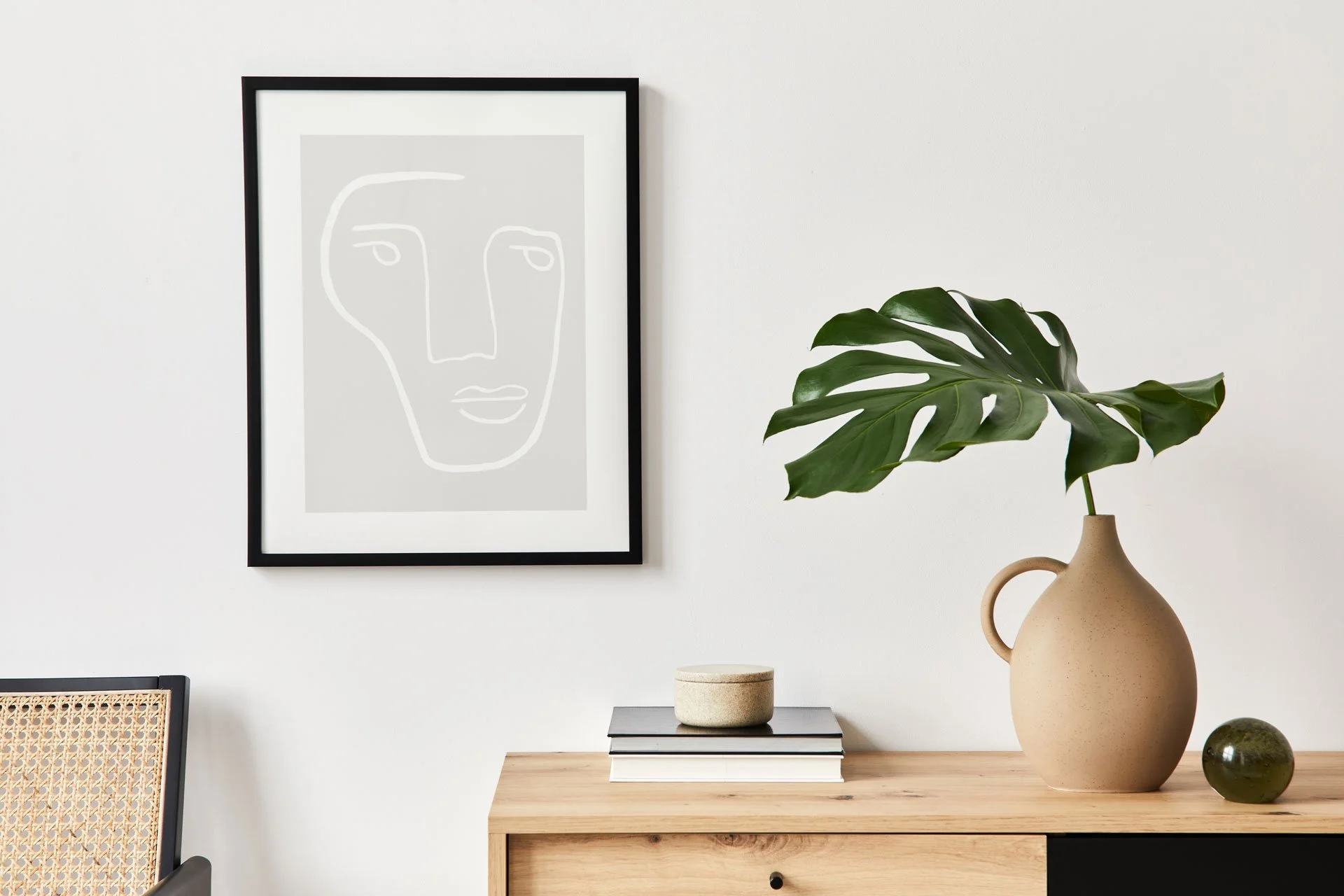 A Minimalist Interior Decor with a monochrome abstract face drawing, a large monstera leaf in a tan ceramic vase, and a small green glass orb on a wooden sideboard.