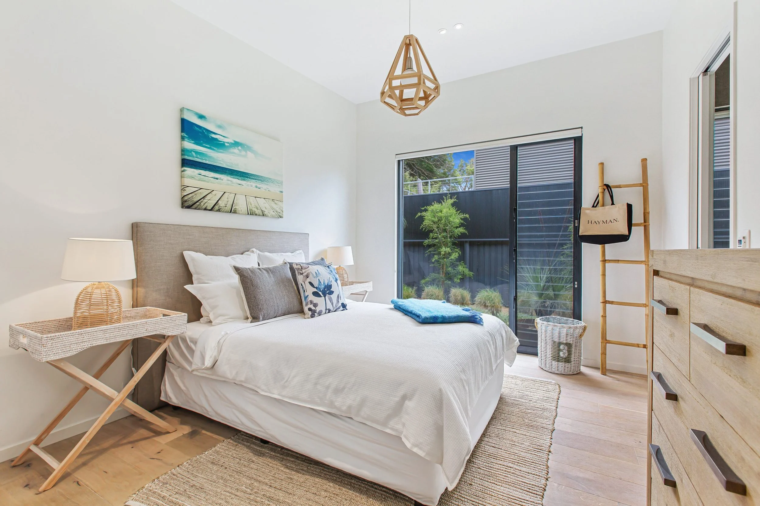 Bright bedroom with white walls, beige upholstered bed, white bedding and decorative pillows, wooden dresser, side tables with lamps, large window with sliding door leading outside, greenery visible through window, wall art of a beach scene, wooden floor with woven rug, wooden ladder with hanging bag, and overhead geometric light fixture.