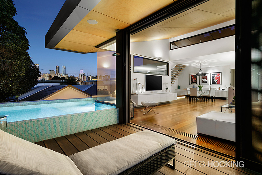 Interior view of a modern apartment with an outdoor pool, living room, and city skyline in the background. The space features a wooden floor, a lounge chair, a TV, a dining table, and artwork on the walls.