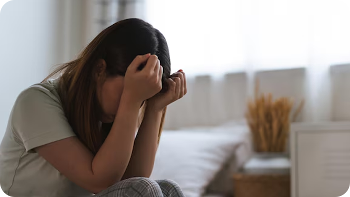 Woman sitting on bed with her head in her hands, appearing upset or distressed in a bright room with a window and dried wheat in the background.