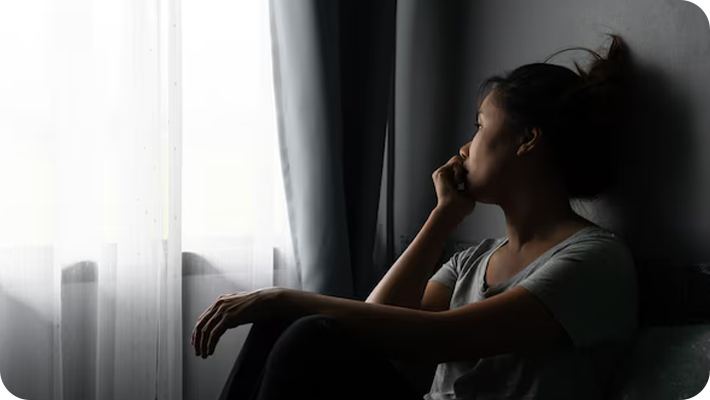 A woman sitting by a window with curtains, gazing outside and resting her chin on her hand.