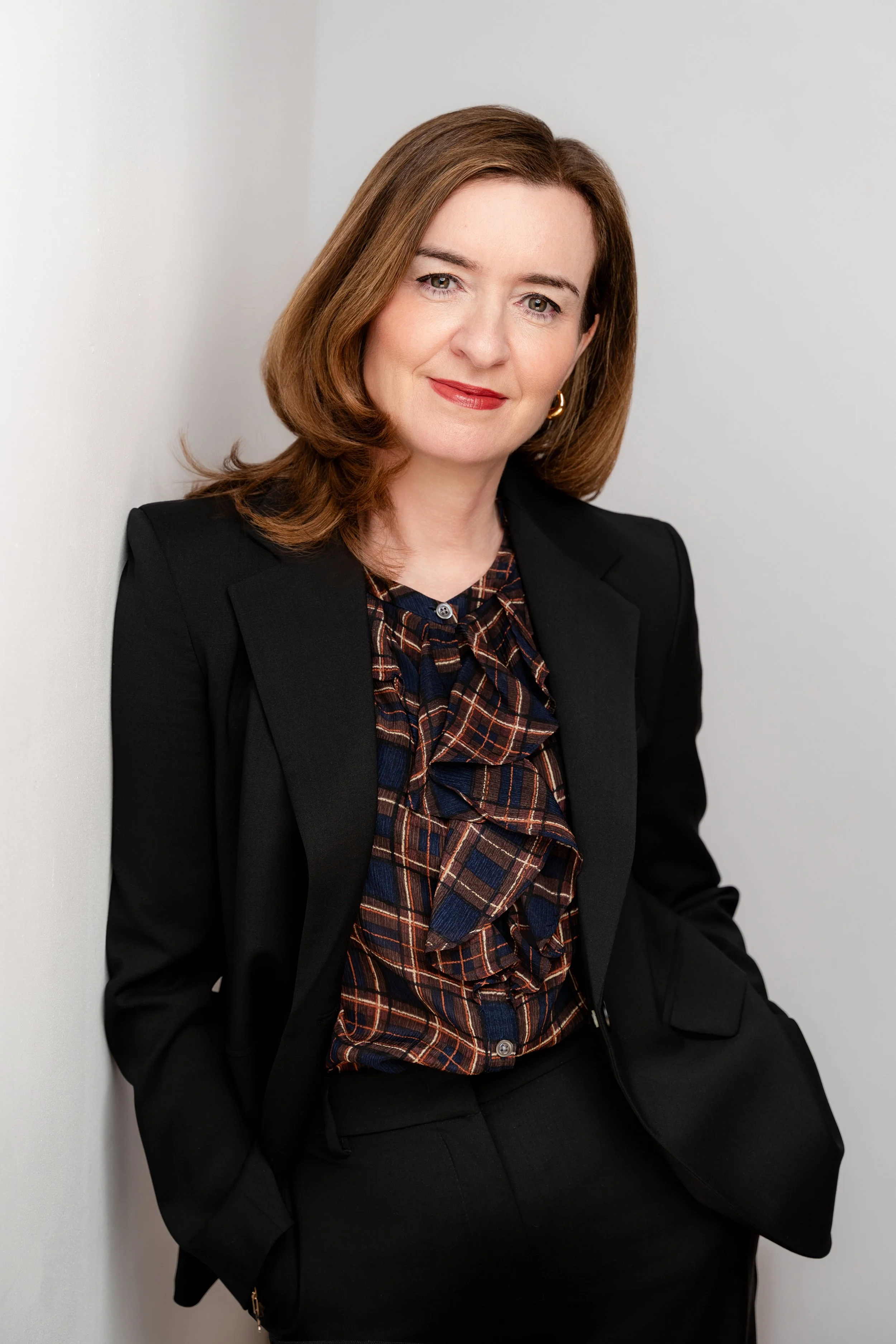 Portrait of a woman with shoulder-length brown hair, wearing a black blazer and a plaid blouse, smiling softly against a plain light-colored background.