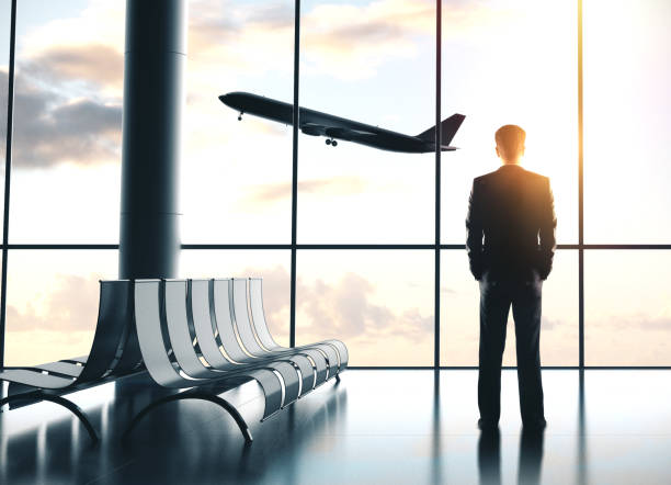 A person standing in an airport terminal, watching an airplane fly outside the large glass windows during sunset.