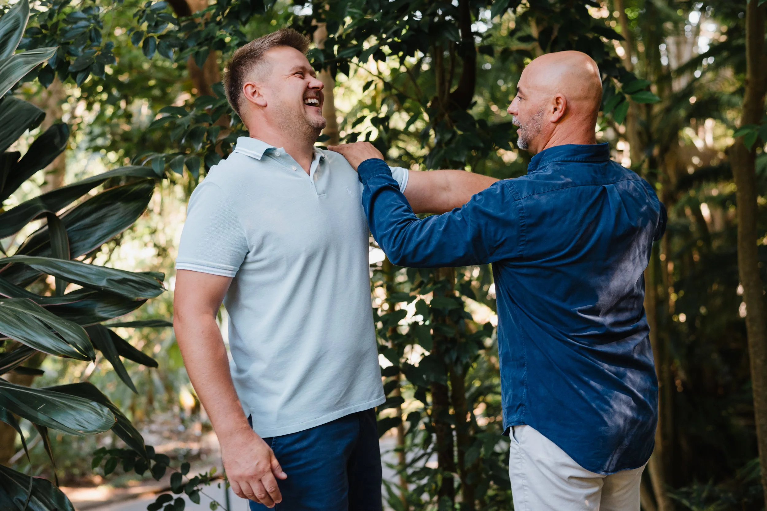 Two men standing outdoors surrounded by greenery, laughing and engaging in conversation.