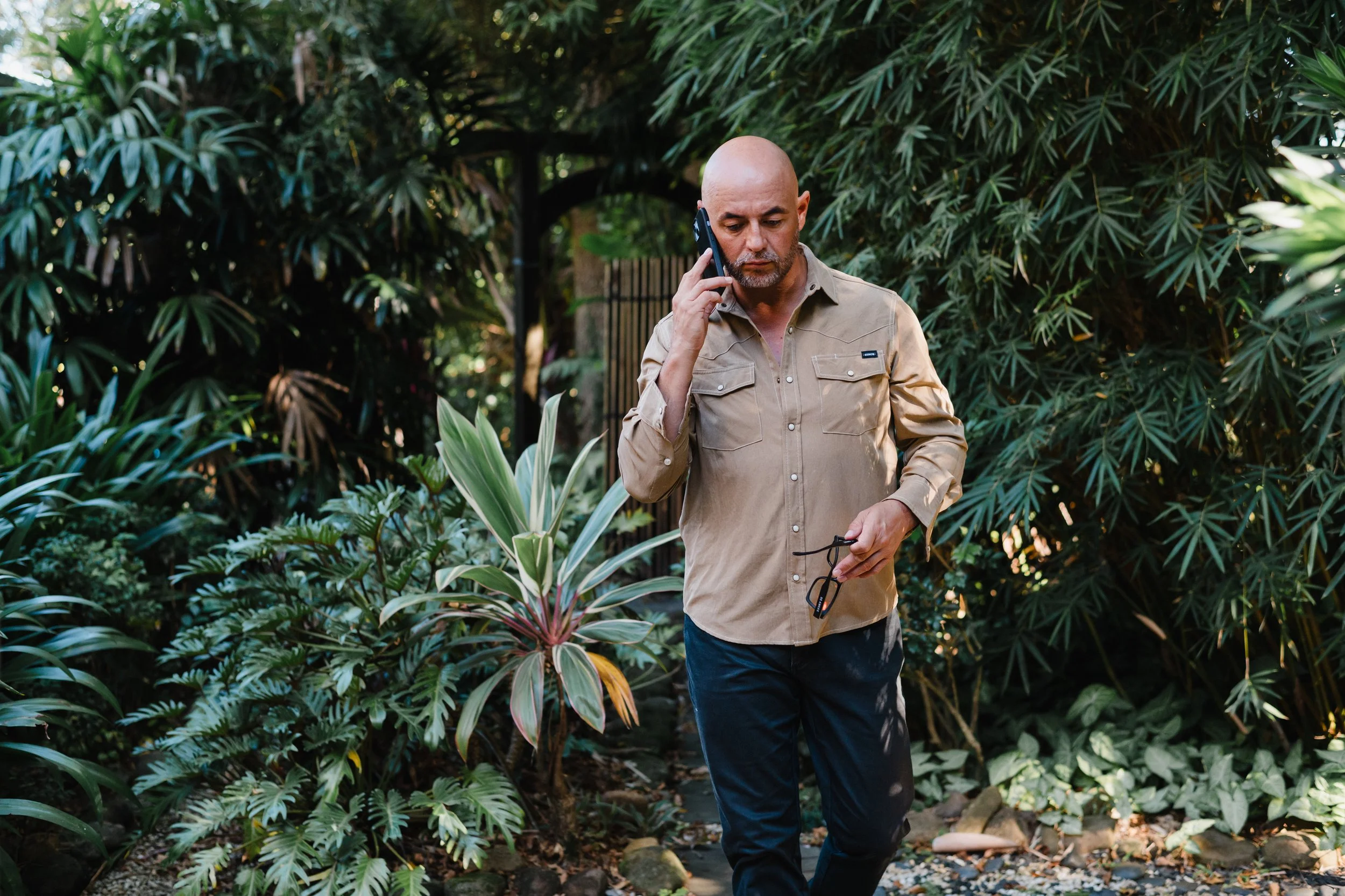 A bald man in a beige shirt standing outdoors among green foliage, talking on his cellphone and holding glasses.