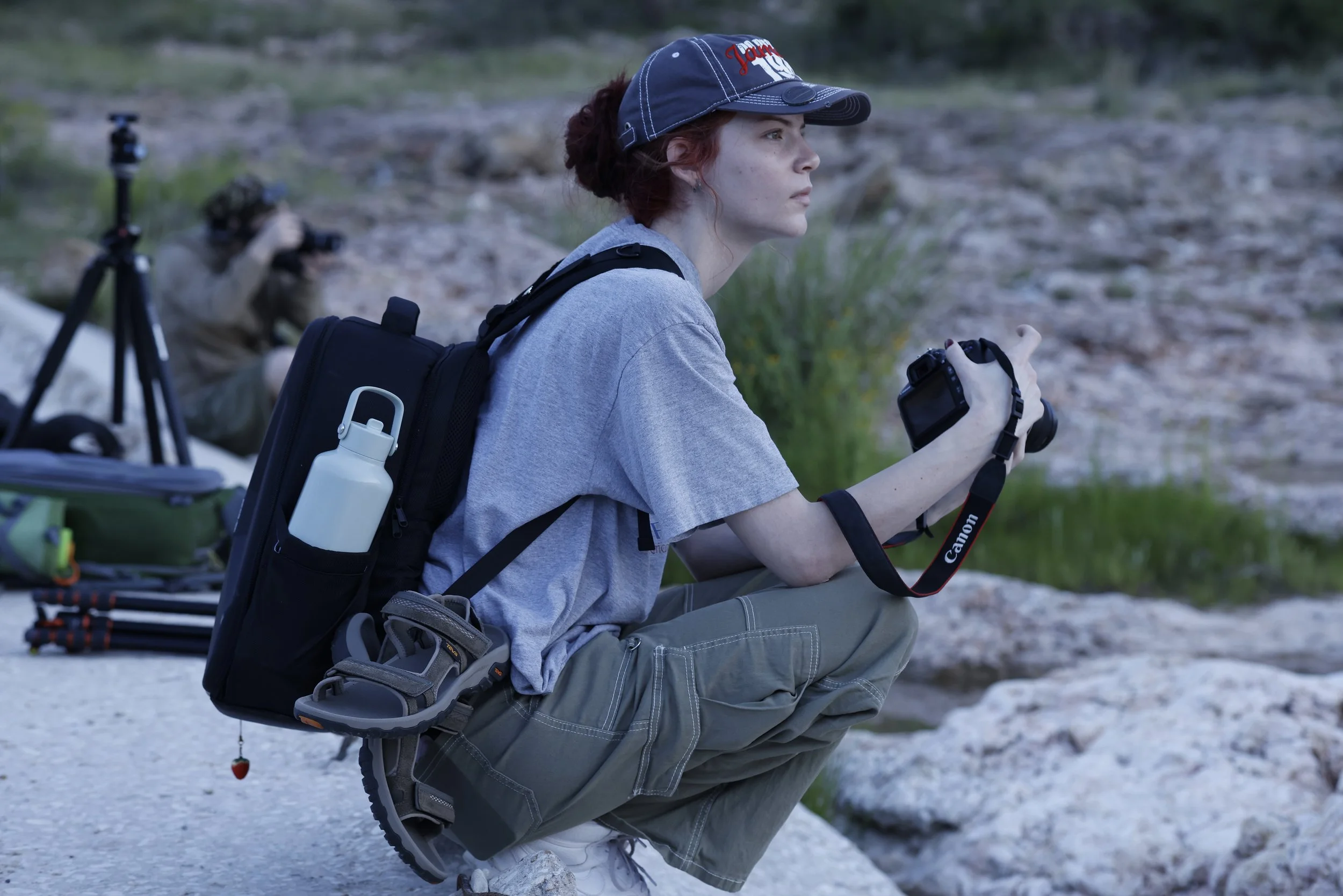 A woman in outdoor clothing kneeling on a rocky surface, holding a camera, with a person in the background adjusting a camera on a tripod; outdoor gear and tripods are around them, suggesting a photography or nature expedition.