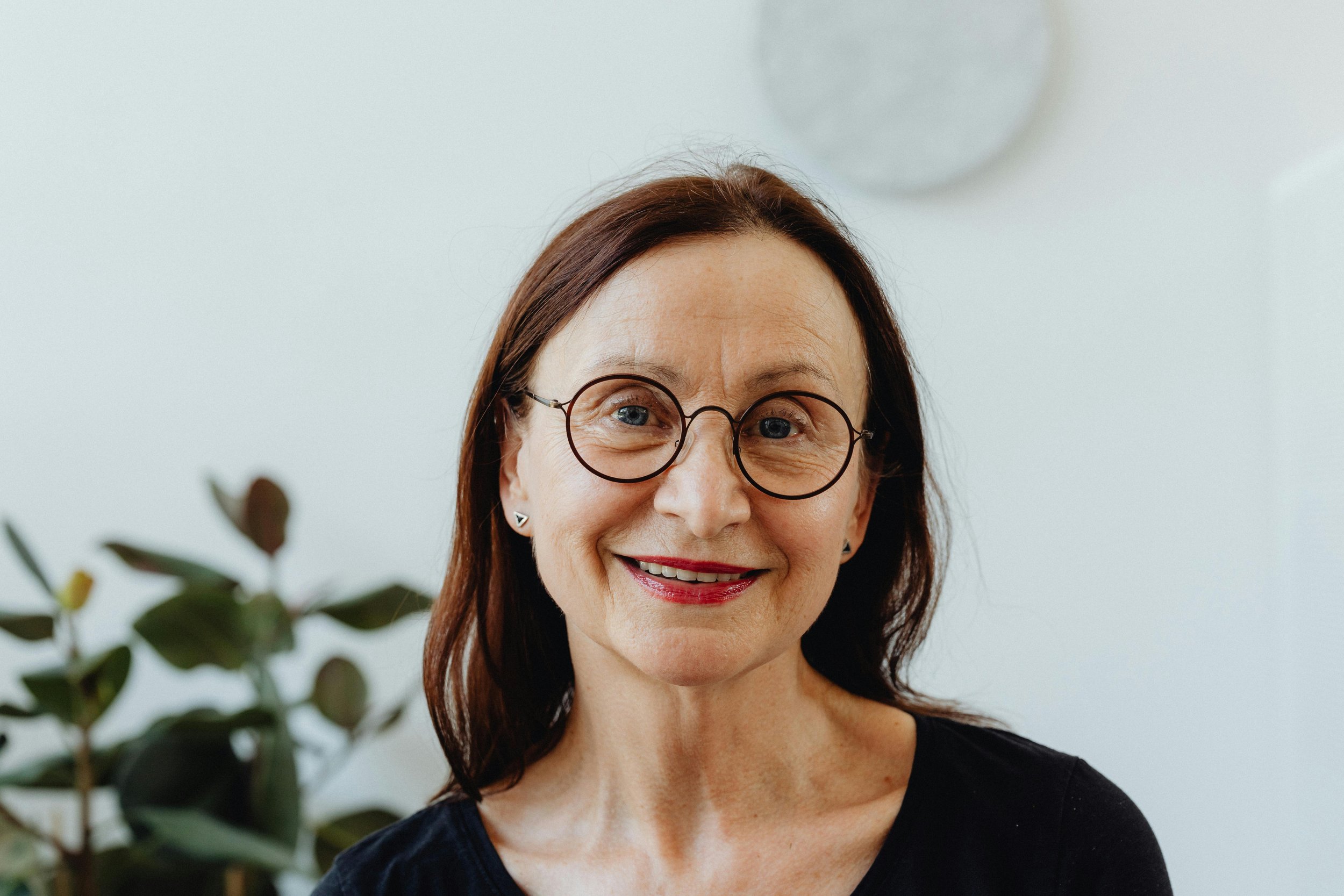 A smiling woman with brown hair, wearing glasses and earrings, standing indoors with a plant in the background.