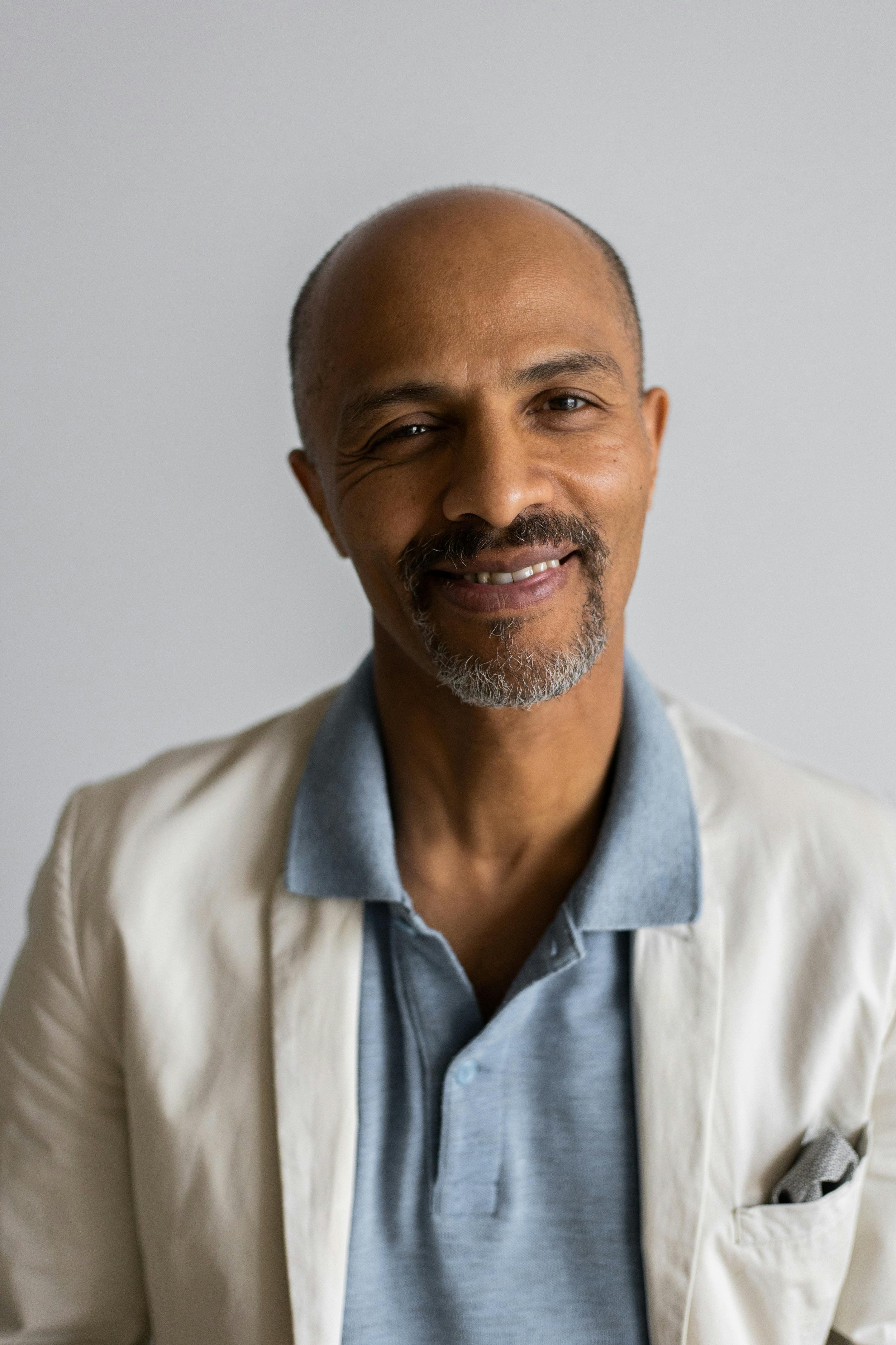 Portrait of a middle-aged man with a bald head, mustache, and goatee, wearing a light beige blazer over a gray collared shirt, smiling against a plain white background.