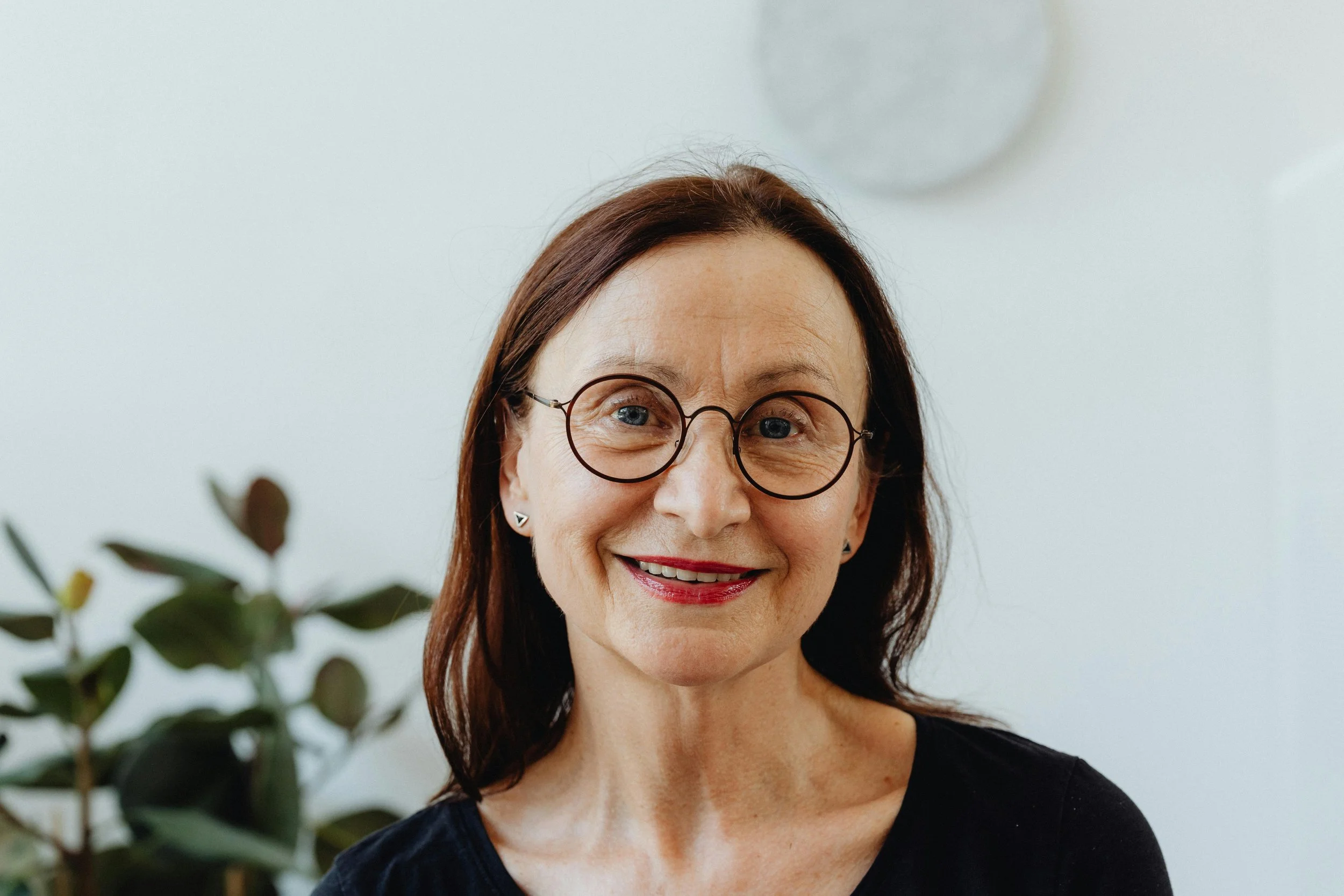 A smiling middle-aged woman with glasses, dark brown hair, and light skin, wearing a black top, standing indoors with a plant and a round clock in the background.
