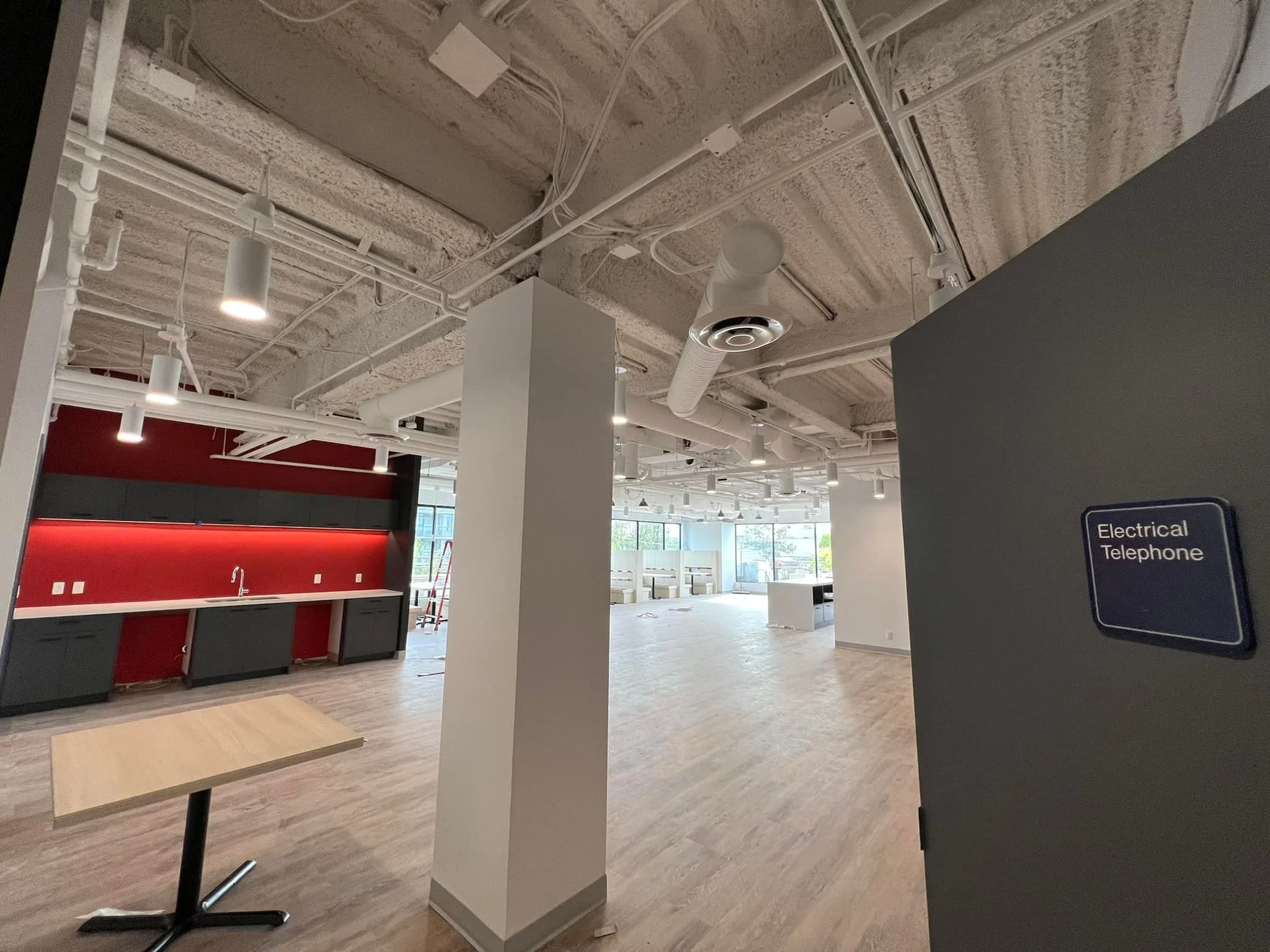 Empty office space with unfinished ceiling, white and gray walls, red and black kitchen area, light wood flooring, and large windows, with a sign on the wall that says 'Electrical Telephone'.